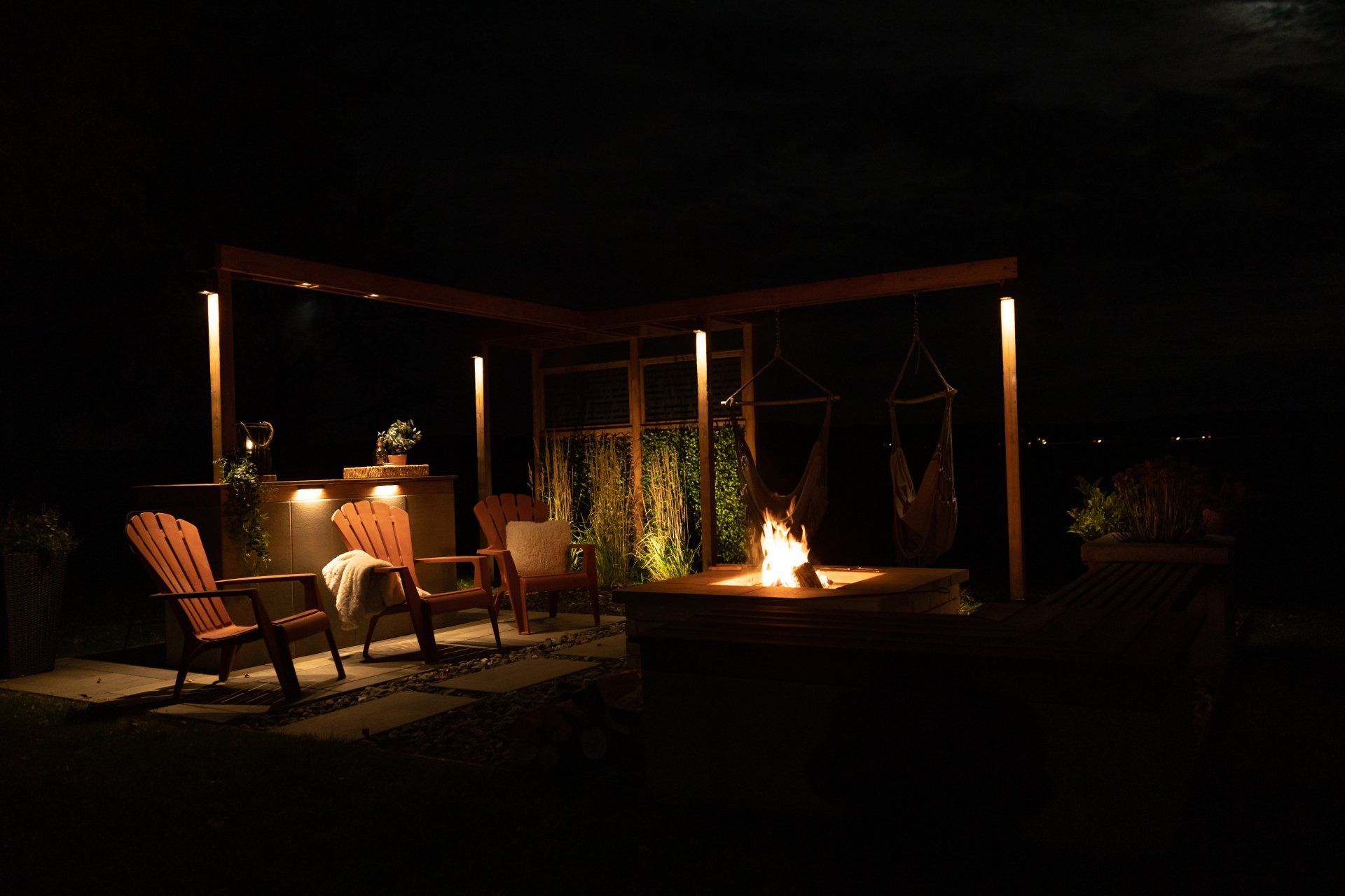 Une terrasse extérieure en soirée avec un foyer, deux chaises en bois, des chaises suspendues et un éclairage d'ambiance chaleureux sur une pergola.