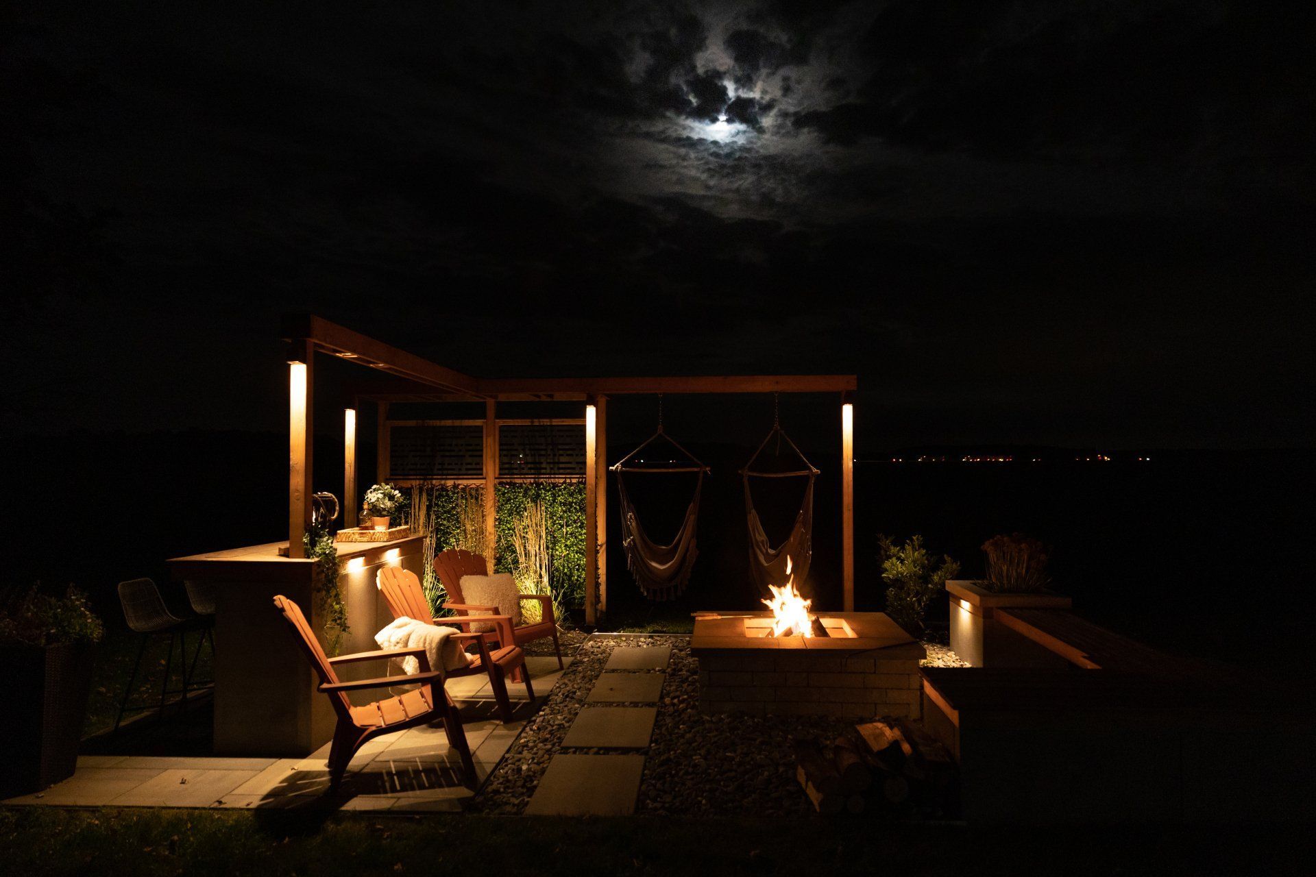 Une terrasse extérieure nocturne avec un foyer, des chaises en bois, un coin bar et des chaises suspendues sous un ciel éclairé par la lune.