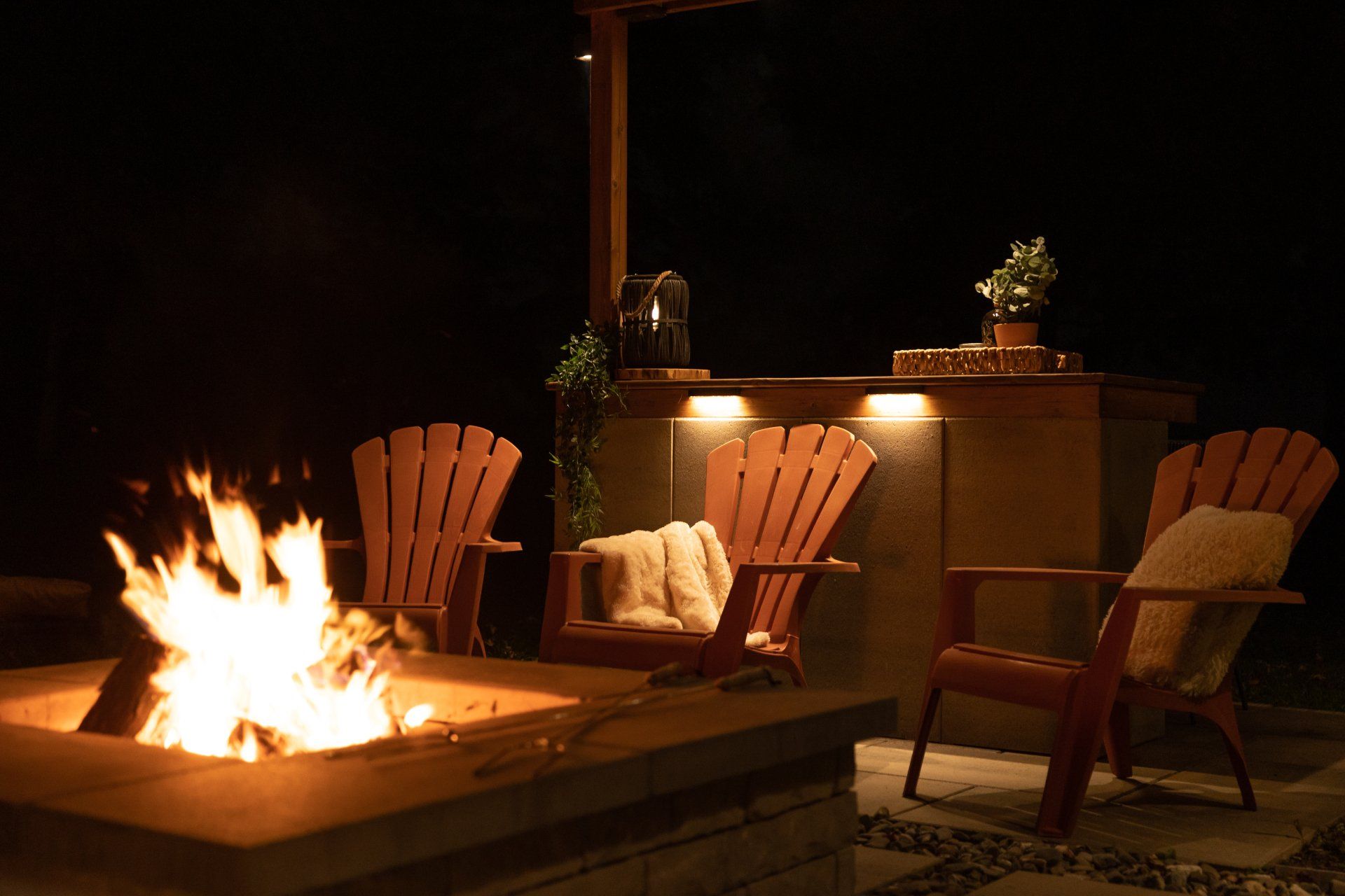 Trois chaises Adirondack disposées autour d'un foyer carré en pierre sur une terrasse, la nuit, avec un bar extérieur éclairé derrière elles.