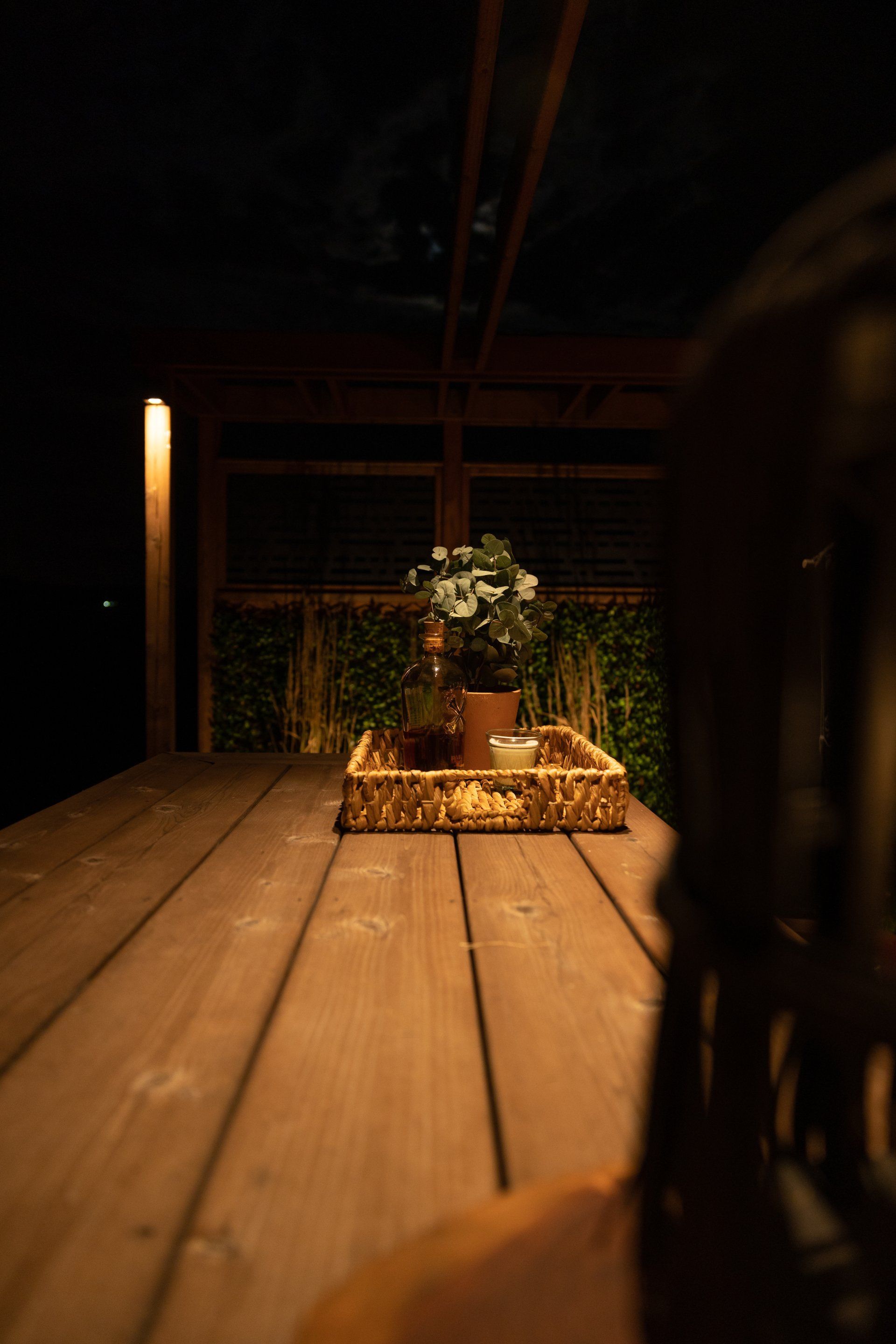 Une table d'extérieur en bois, la nuit, avec une petite plante en pot dans un plateau tressé, éclairée par une lumière chaude et tamisée.