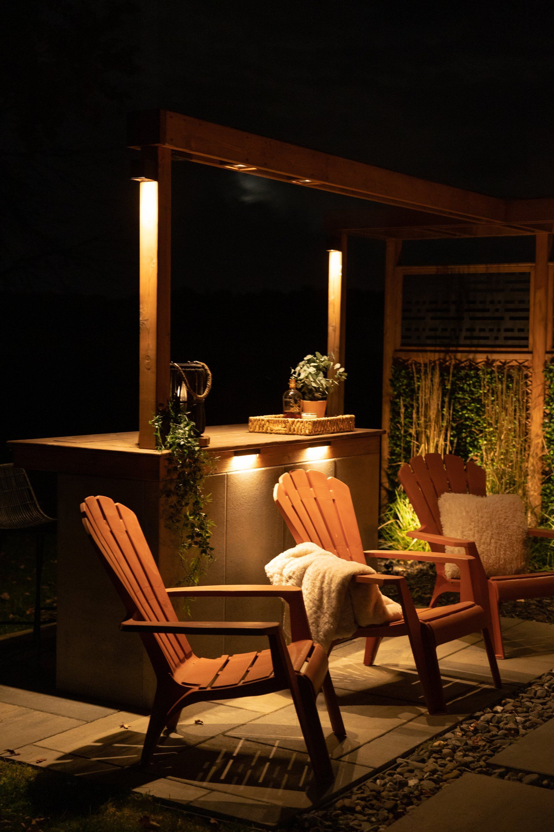 Deux chaises Adirondack en bois font face à un bar de jardin illuminé par des guirlandes lumineuses aux tons chauds, sur une terrasse en soirée.