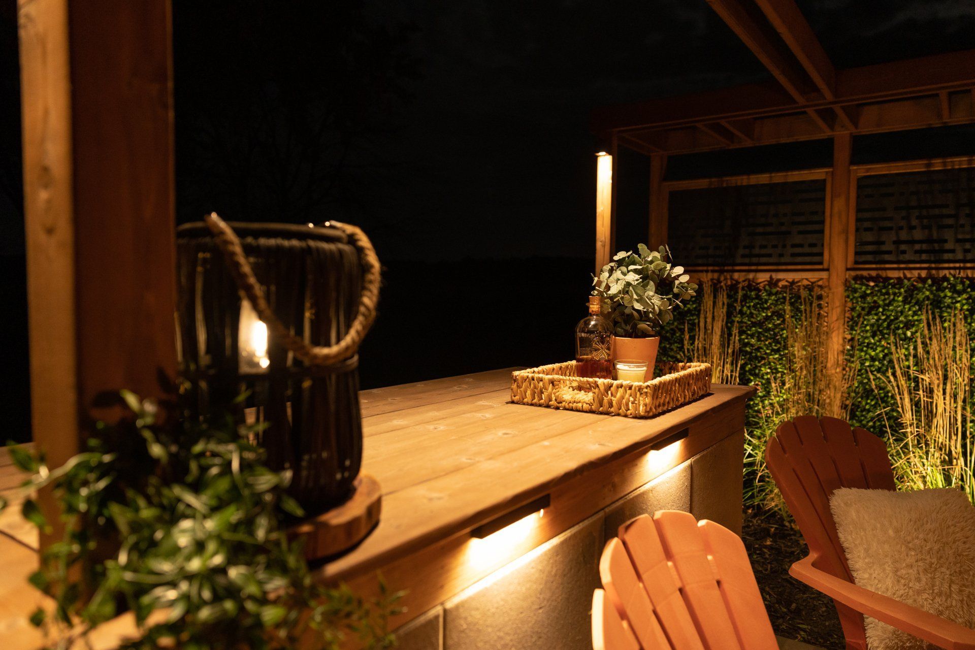 Un bar extérieur en bois, la nuit, éclairé par une lanterne et des lumières chaudes sous le comptoir, avec des chaises et un plateau de boissons.