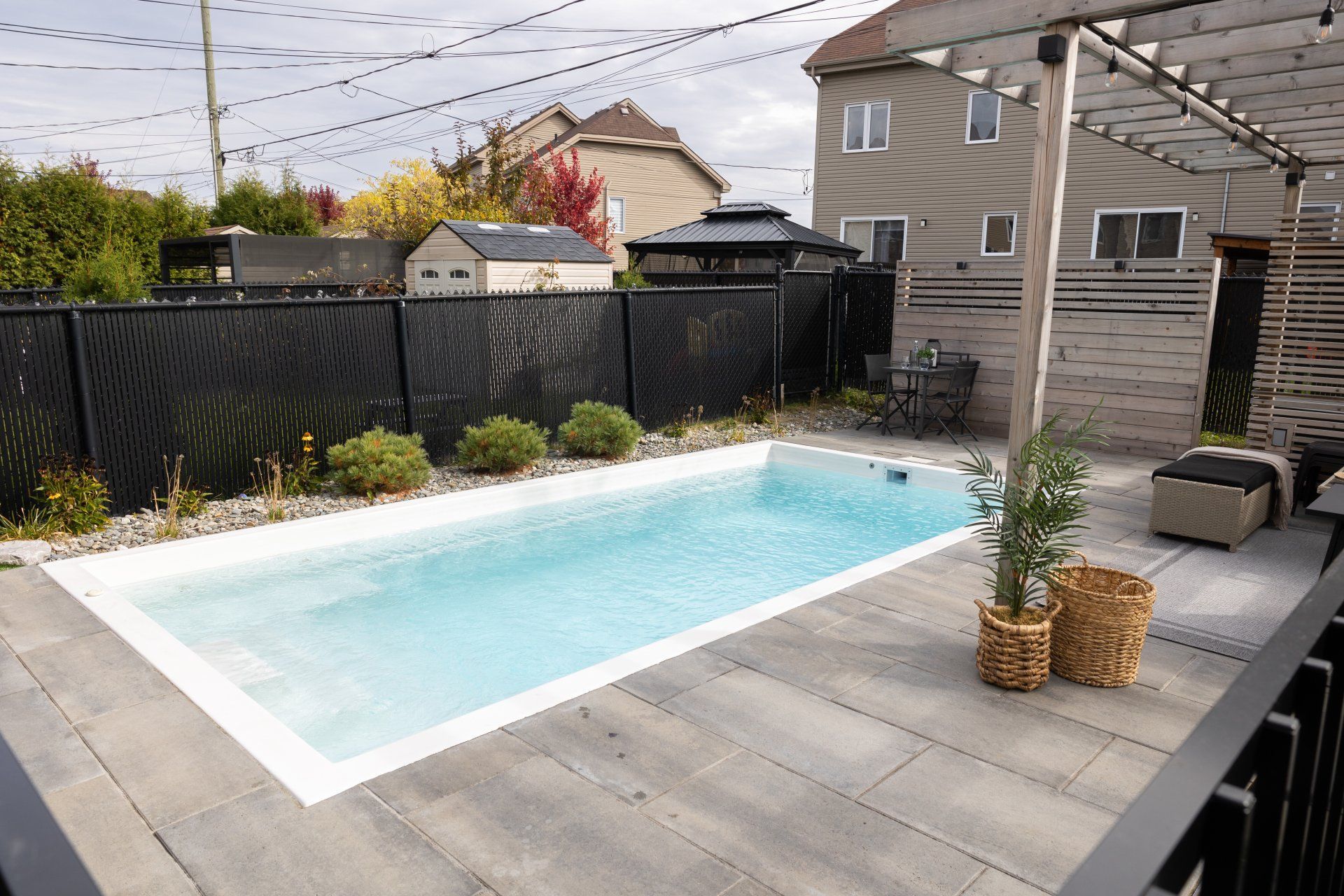 Une piscine rectangulaire entourée d'une terrasse en pierre grise, d'une clôture noire pour préserver l'intimité et d'une pergola en bois dans un jardin.
