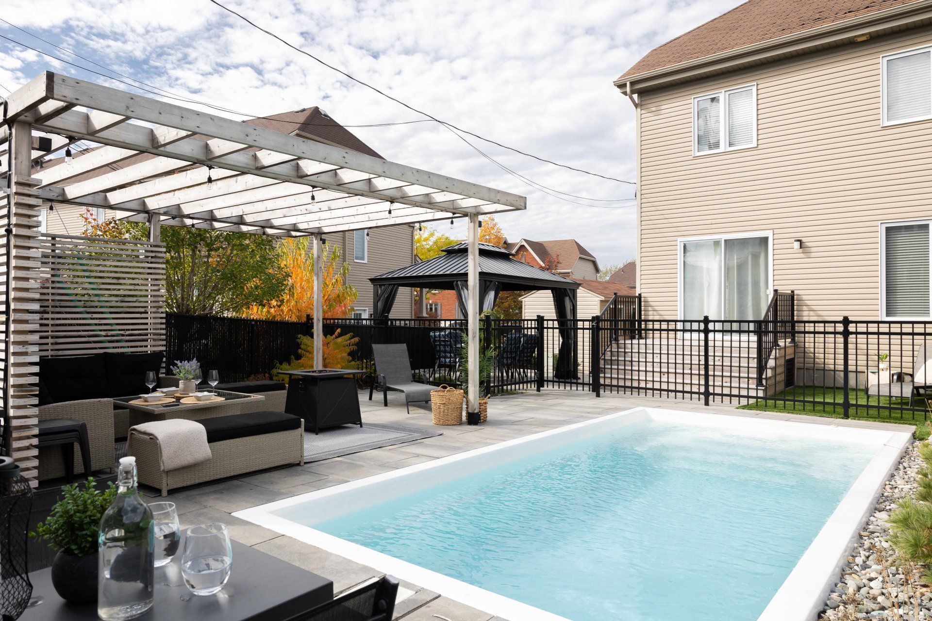 Un espace piscine moderne dans le jardin, avec une terrasse pavée de couleur claire, un coin détente sous une pergola et une maison beige à deux étages.