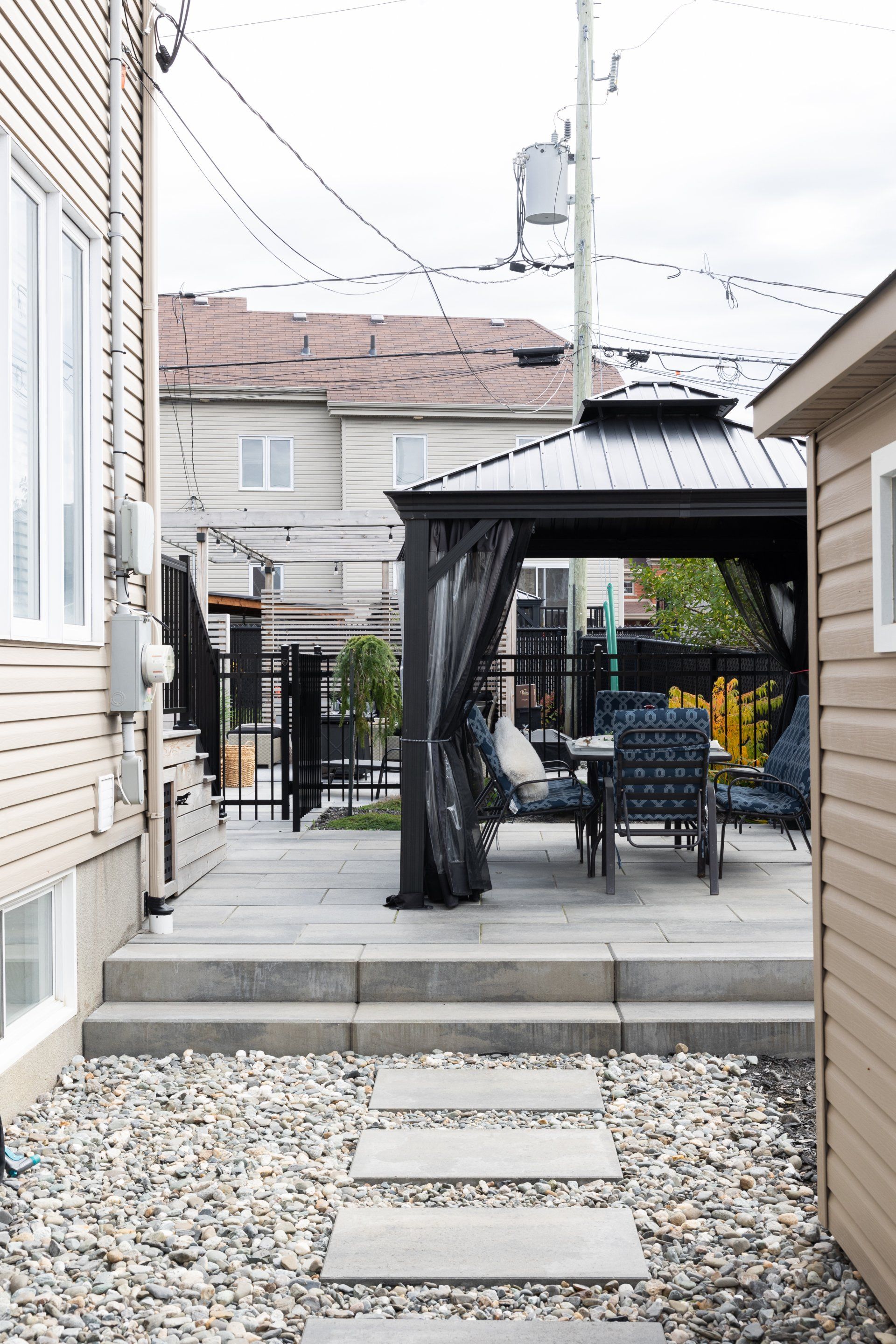 Une terrasse avec un kiosque sombre, des chaises de salle à manger et un chemin de pierre menant à des marches en béton entre deux murs de bardage beige.