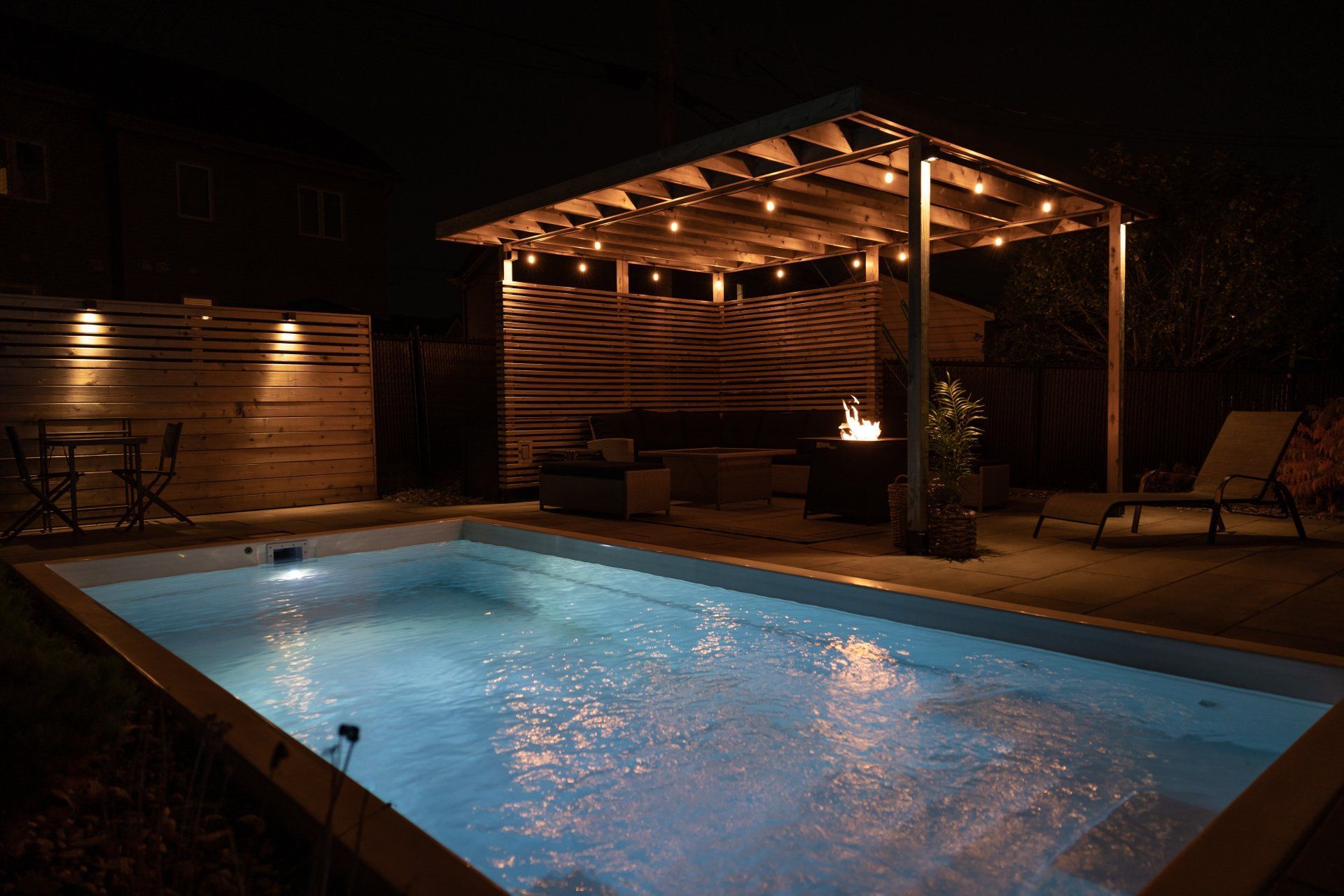 Une piscine illuminée dans le jardin la nuit, avec une pergola couverte ornée de guirlandes lumineuses, un foyer et un coin salon confortable sur la terrasse.