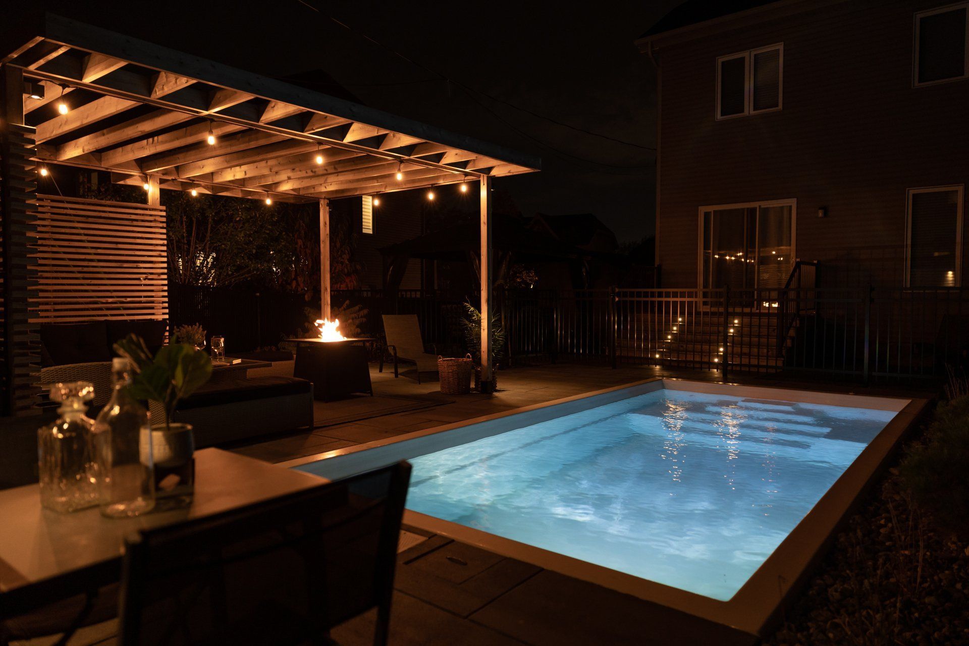 Une piscine illuminée la nuit, jouxtant une terrasse ornée de guirlandes lumineuses, d'un foyer et de sièges sous une pergola en bois.