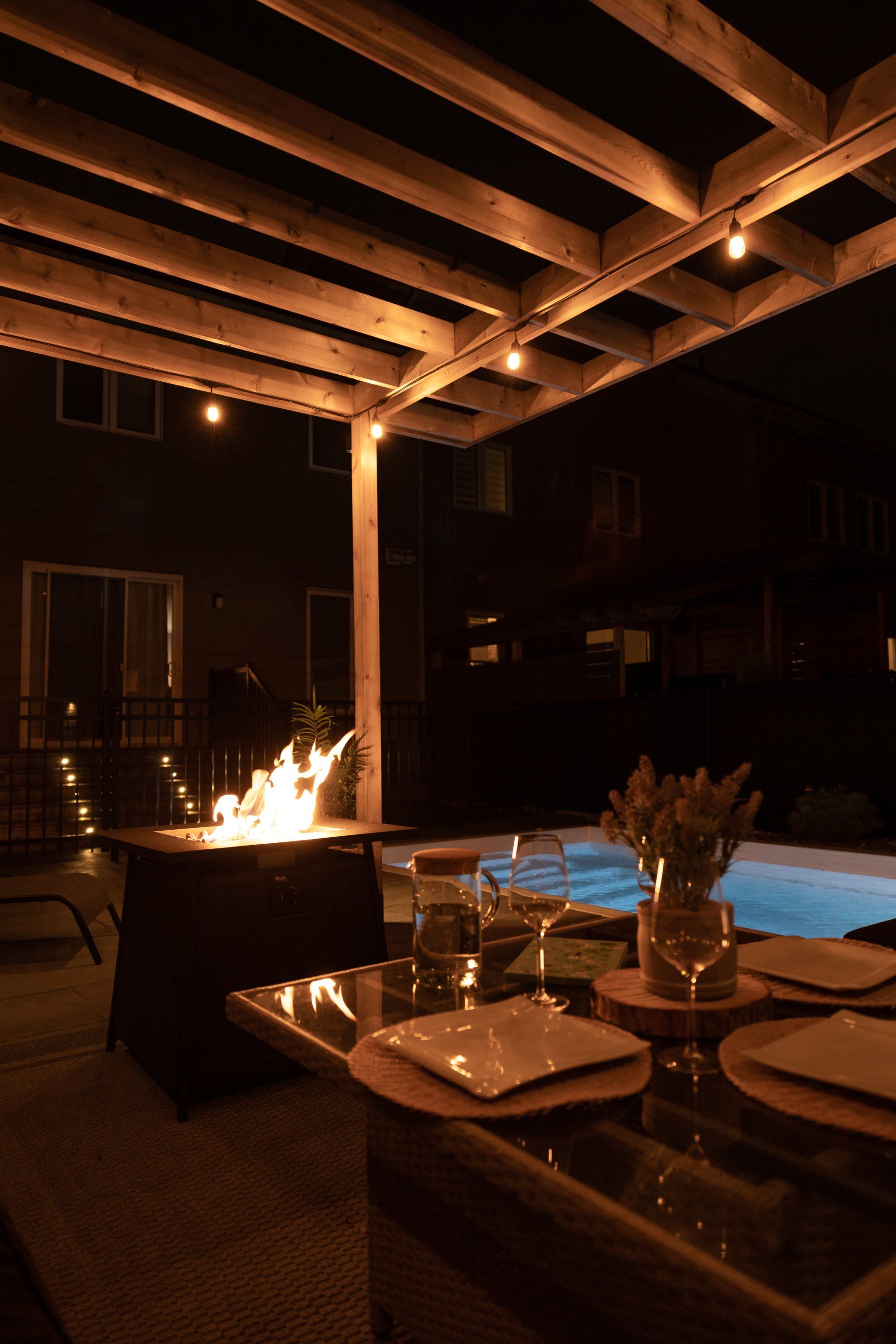 Un foyer allumé sur une terrasse le soir, avec une piscine et une table à manger dressée sous une pergola en bois illuminée de guirlandes lumineuses.