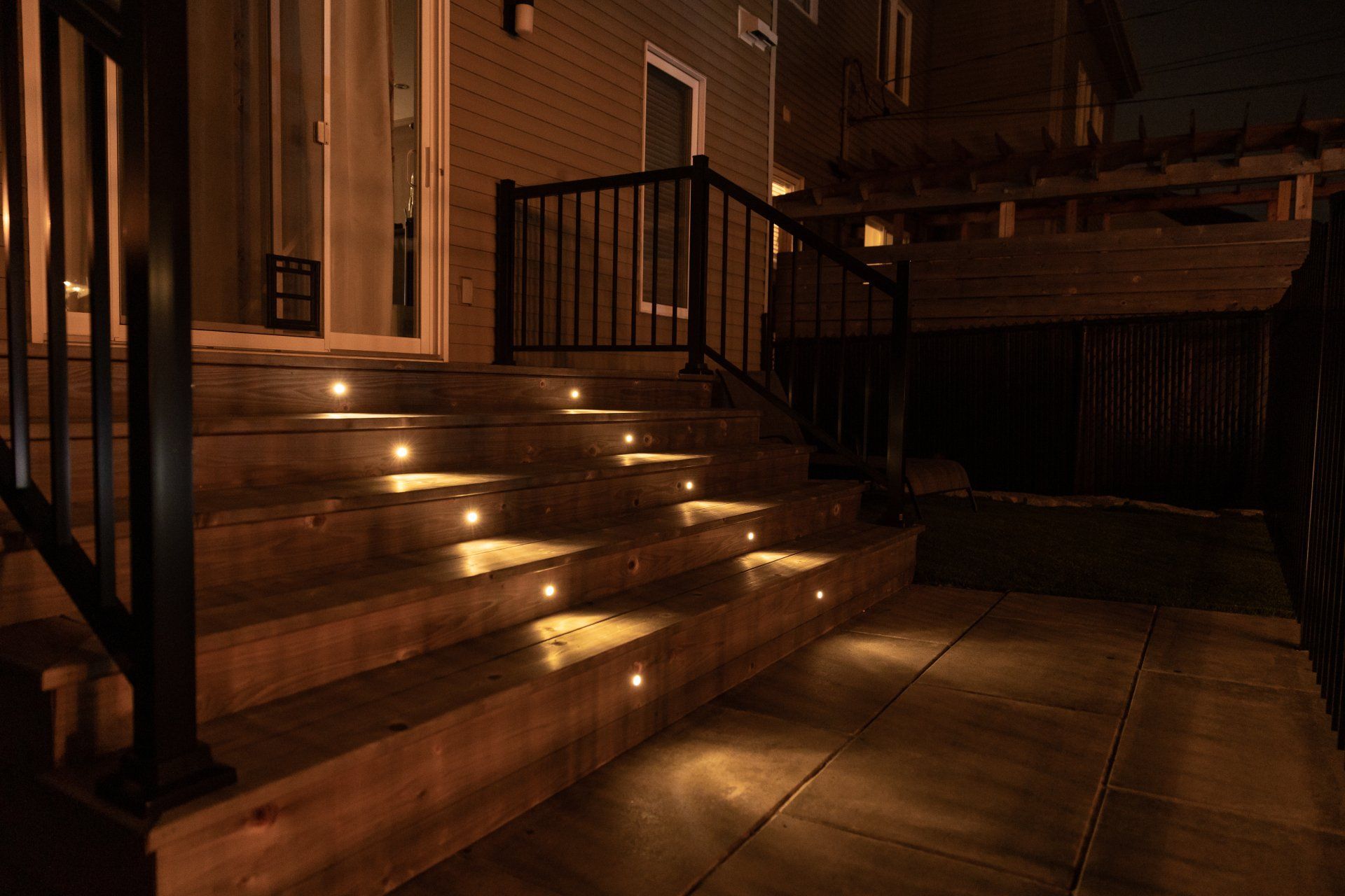 Vue nocturne de marches de terrasse en bois illuminées par de petites lumières circulaires à la lumière chaude, menant au patio d'une maison.