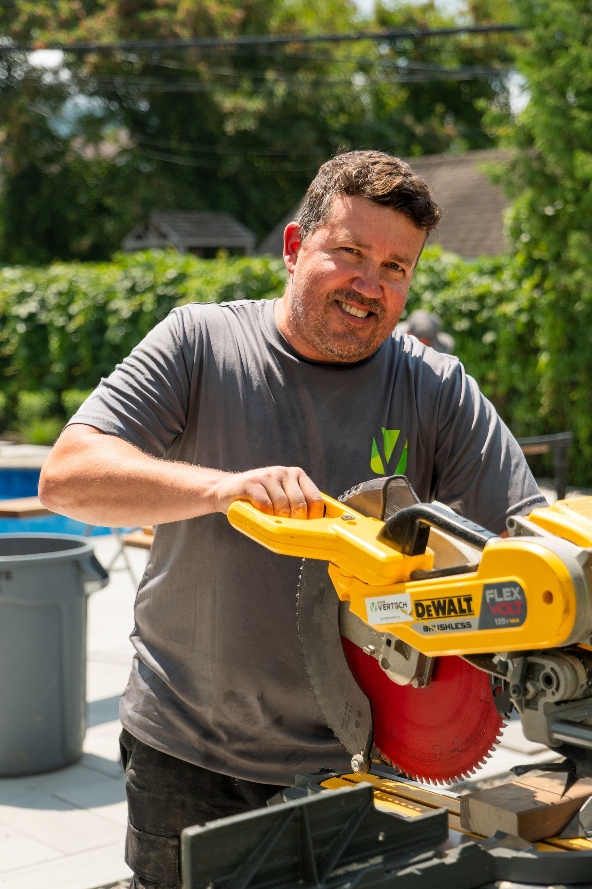 Une personne souriante utilise une scie à onglets DeWalt jaune à l'extérieur, dans un jardin ensoleillé et verdoyant.