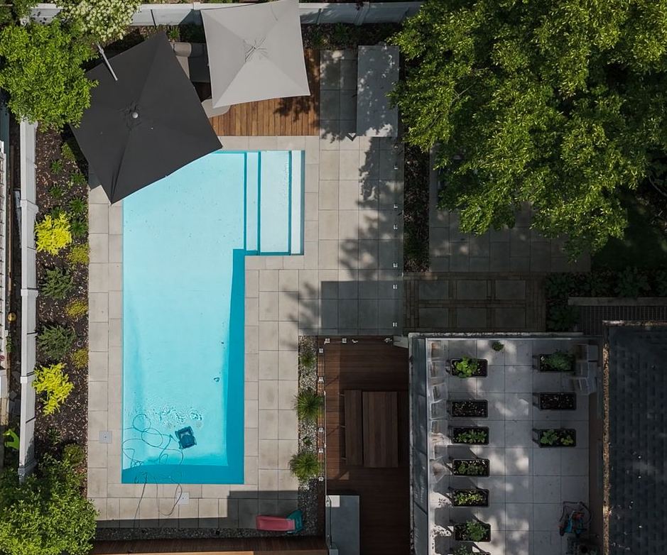 Vue aérienne d'une piscine rectangulaire dans un jardin avec un robot nettoyeur, une terrasse en pierre adjacente, une terrasse en bois et un espace jardin.