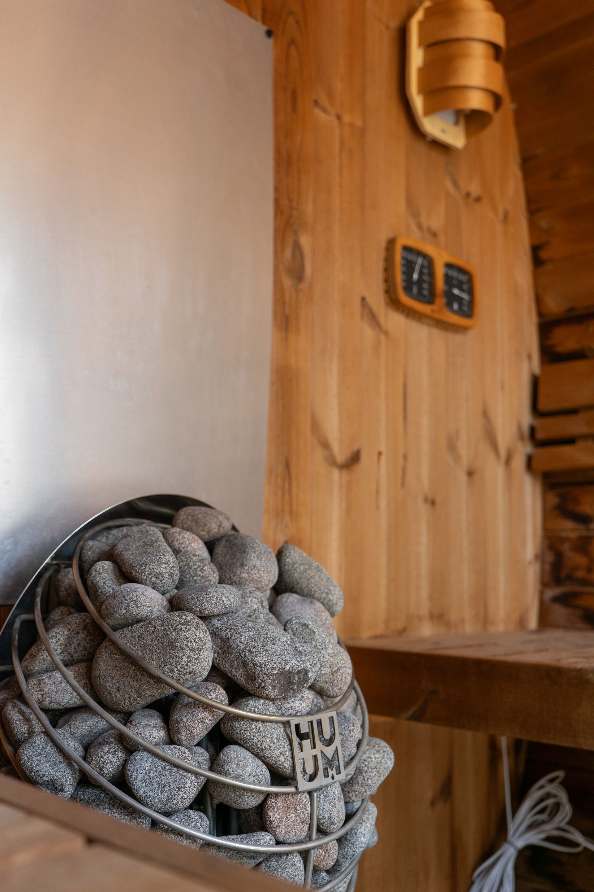 Un poêle de sauna rempli de pierres grises est placé devant des murs lambrissés, avec une lampe murale et un thermomètre.