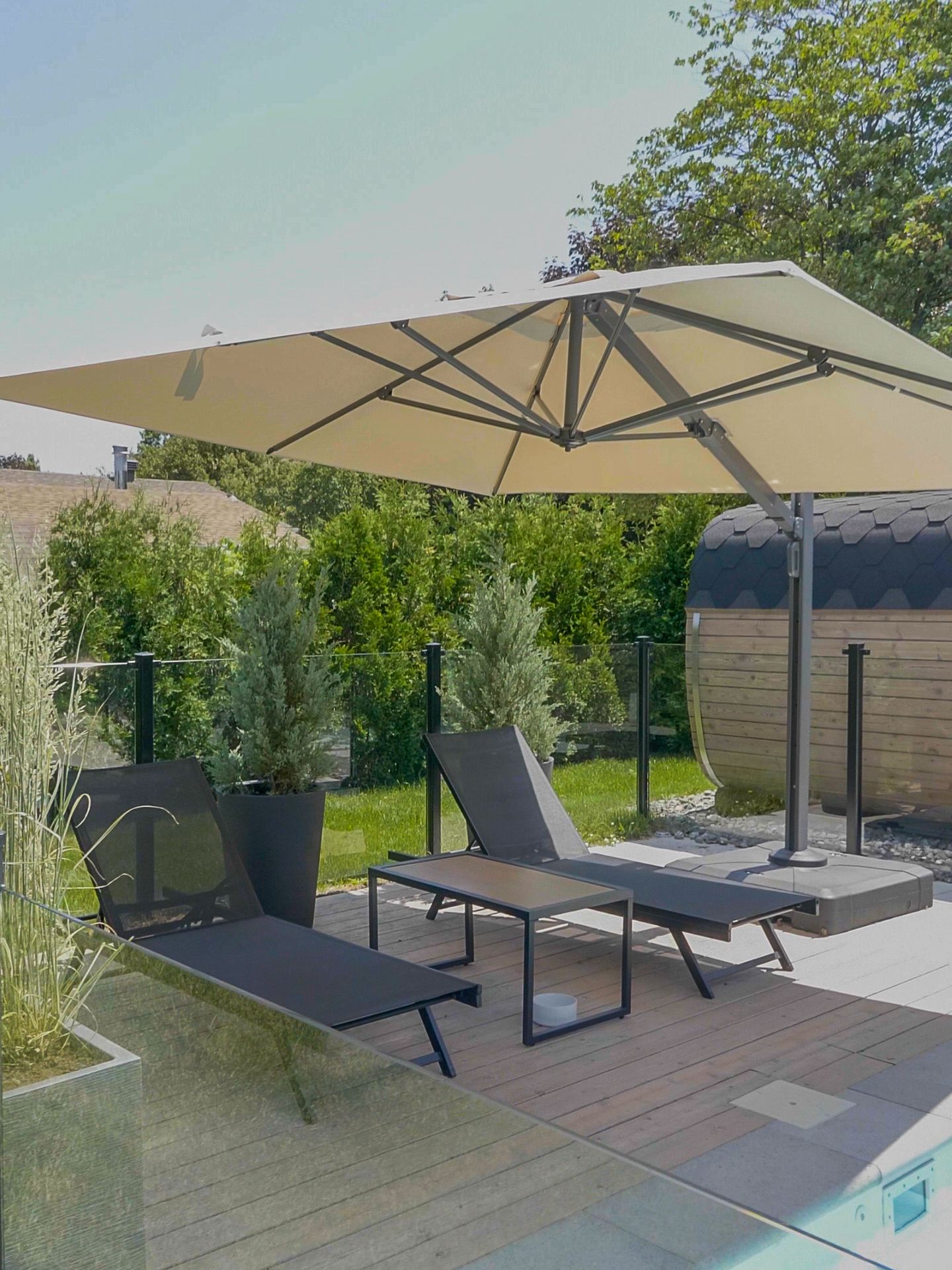 Deux chaises longues et une petite table sous un grand parasol beige déporté sur une terrasse ensoleillée au bord d'une piscine.