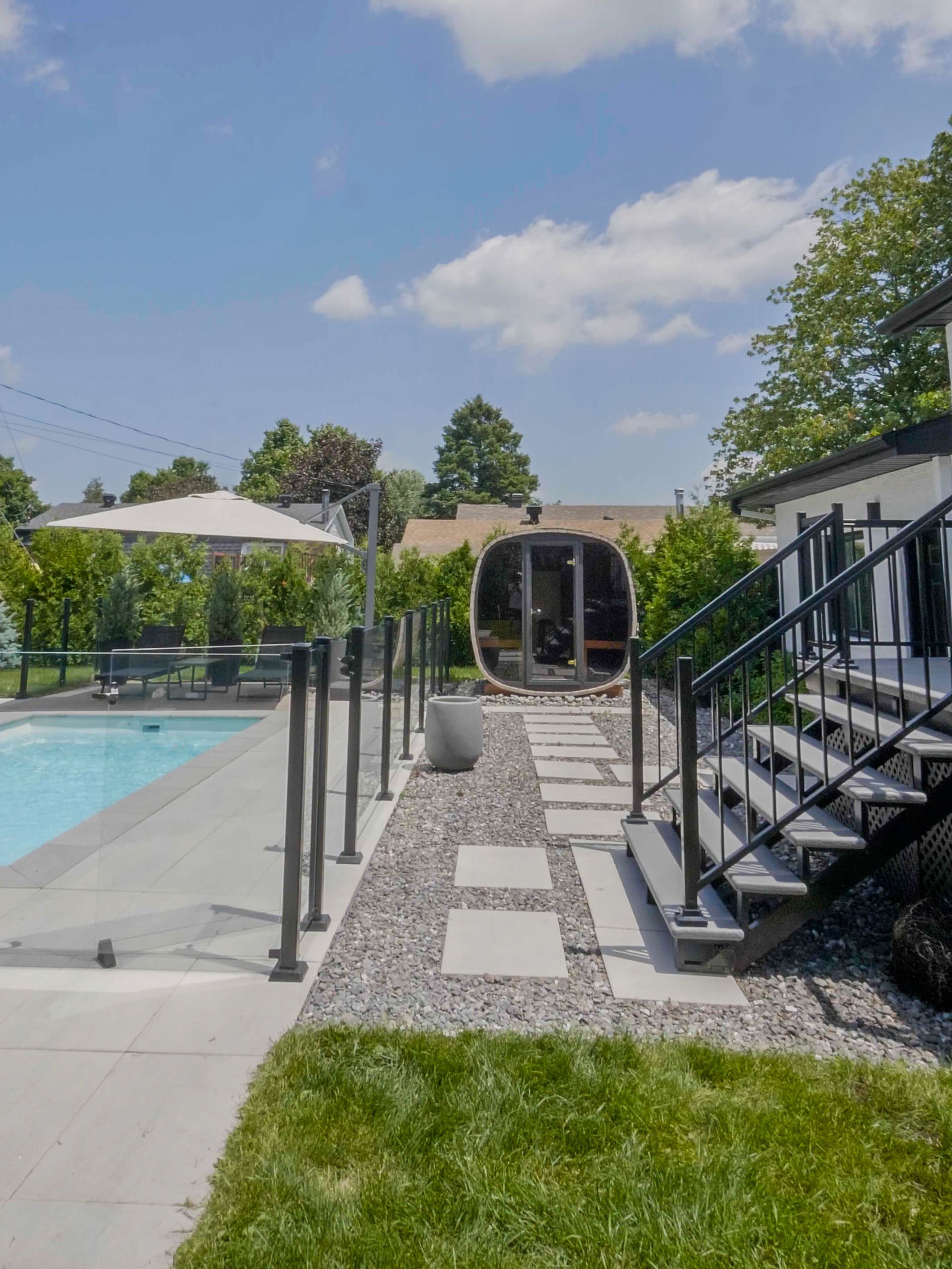 Une terrasse dans le jardin avec piscine, clôture en verre, allée de gravier avec pavés et sauna en forme de tonneau sous un ciel bleu azur.