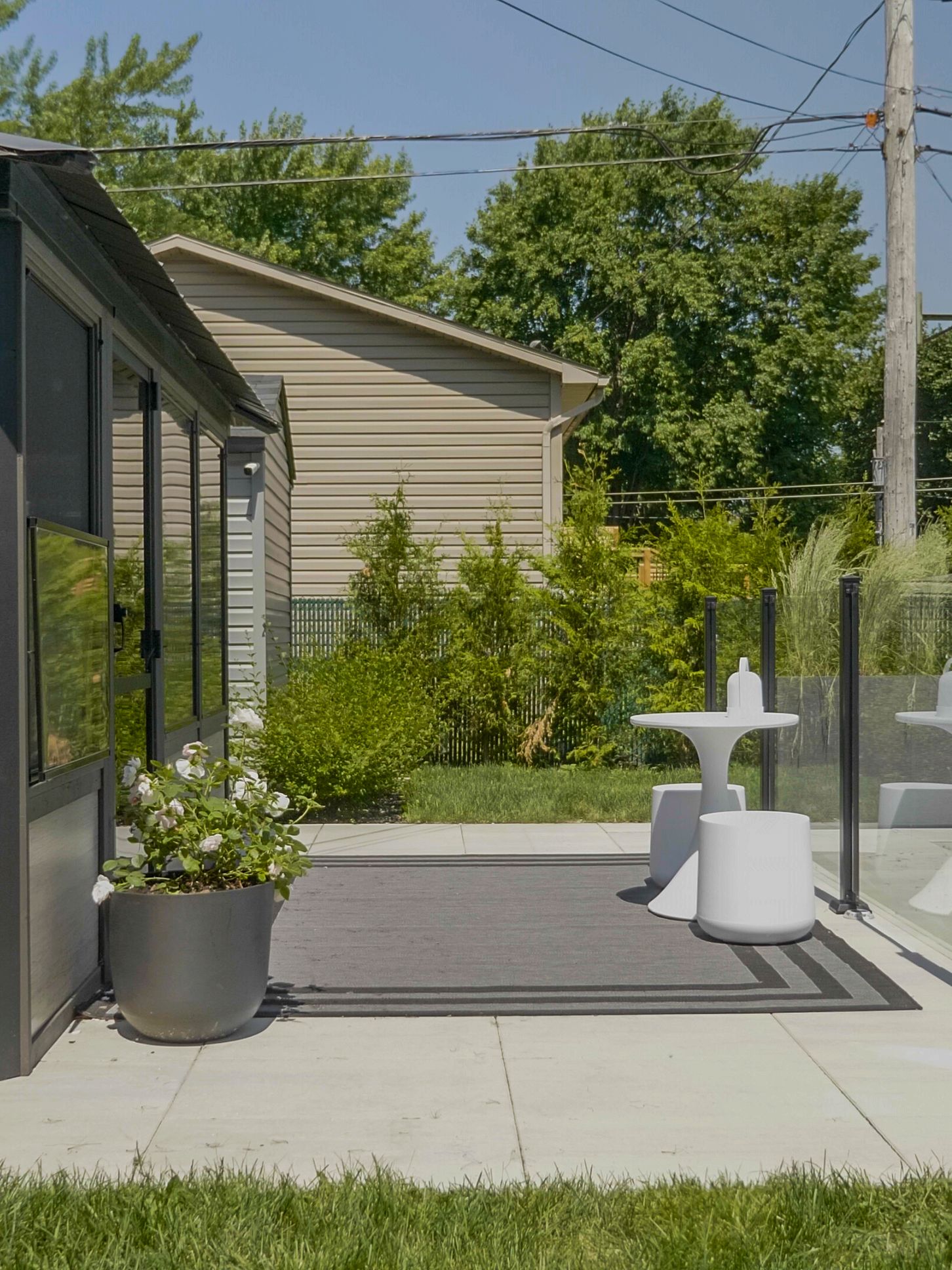 Une terrasse avec un tapis gris foncé, une plante en pot et une chaise bistro blanche installée à côté d'une structure moderne protégée par un écran, par une journée ensoleillée.