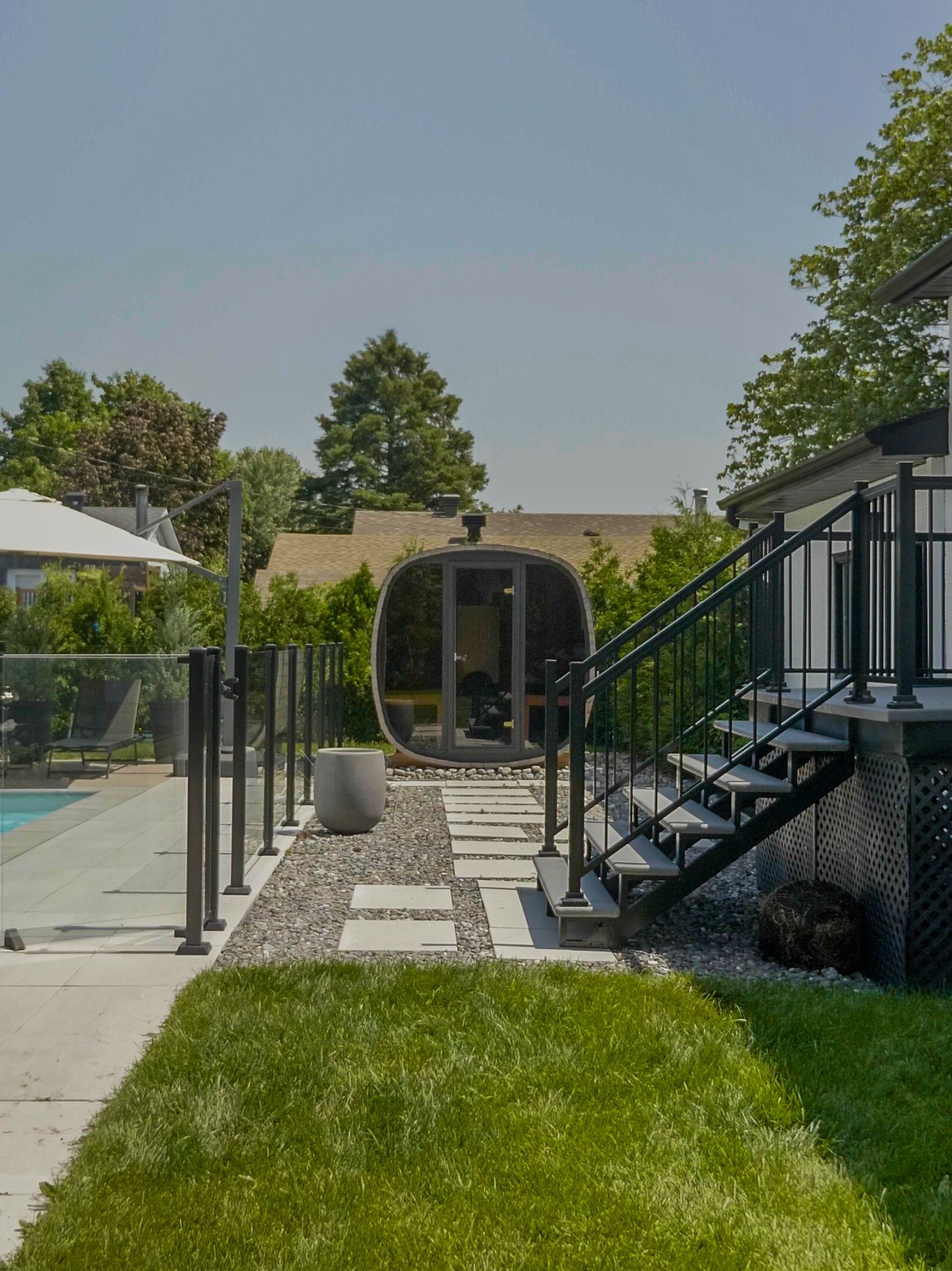 Vue d'un jardin montrant un sauna en forme de tonneau sur un chemin de pierre, à côté d'une piscine, d'une pelouse et d'un escalier.