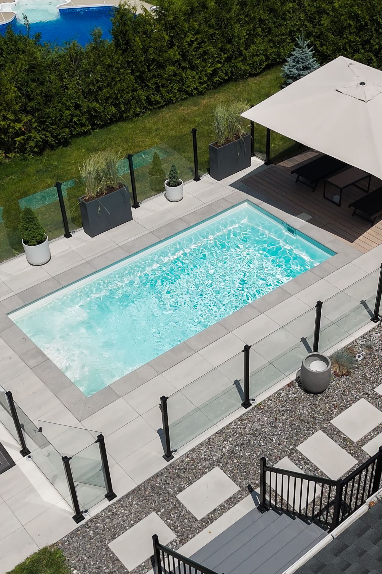 Une piscine rectangulaire dans le jardin, entourée de dalles de pierre gris clair, d'une clôture en verre et d'une terrasse ombragée.