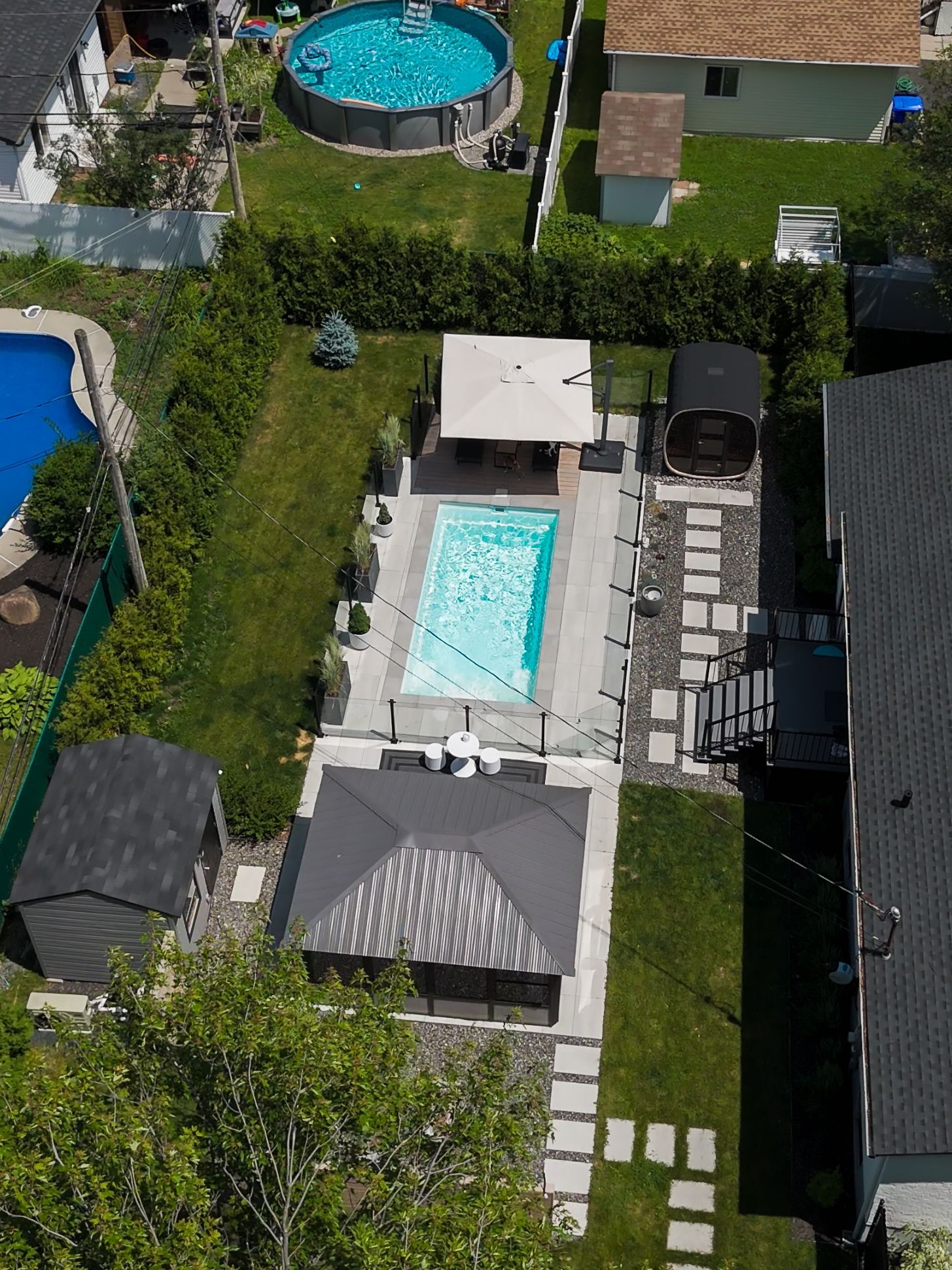 Vue aérienne d'un jardin comprenant une piscine rectangulaire, un grand kiosque, un petit abri de jardin et une allée de terrasse en pierre.