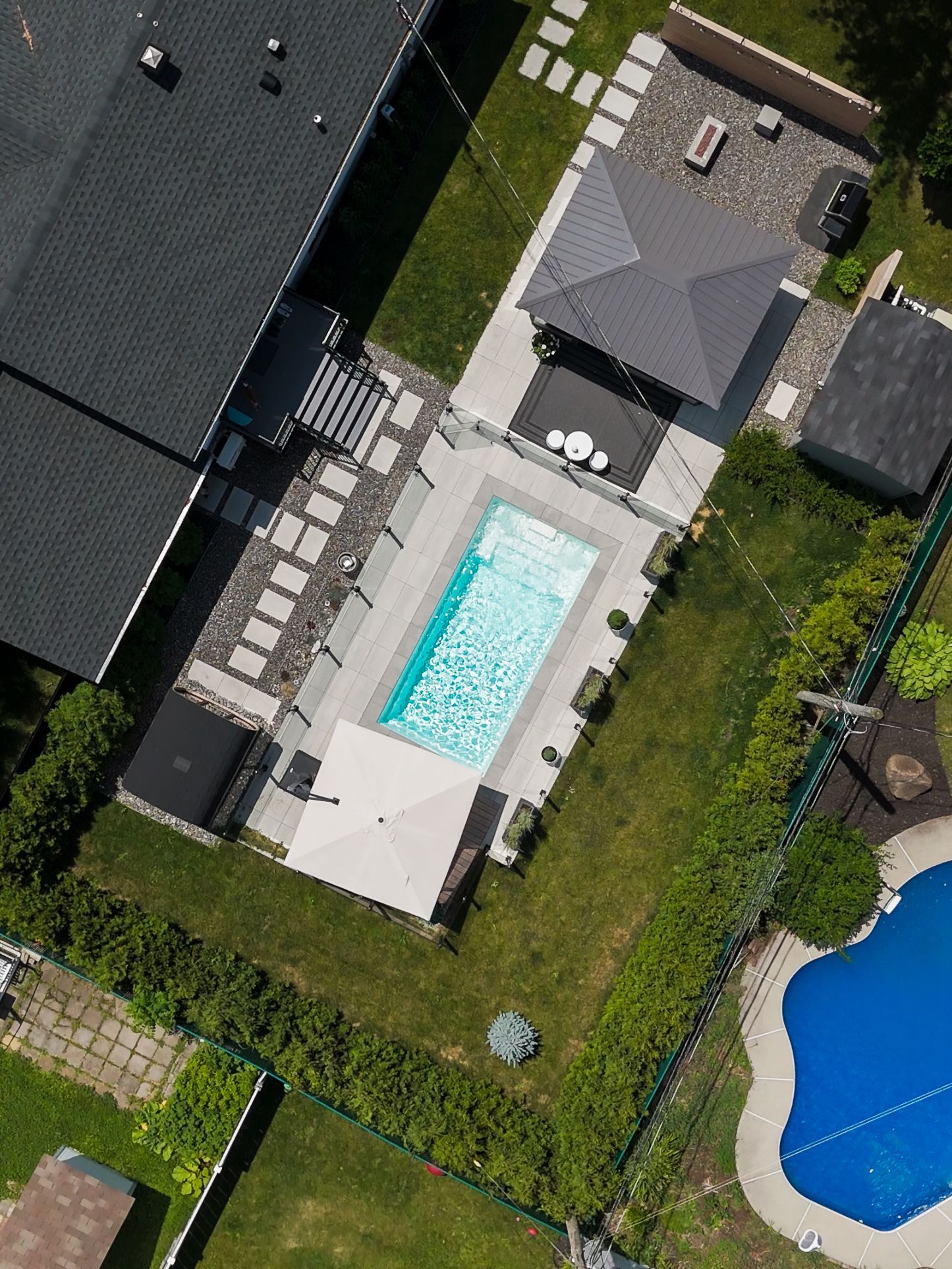 Vue aérienne d'une piscine rectangulaire dans un jardin, entourée d'une terrasse en pierre claire, d'un kiosque et d'une piscine bleue en forme de rein située à proximité.
