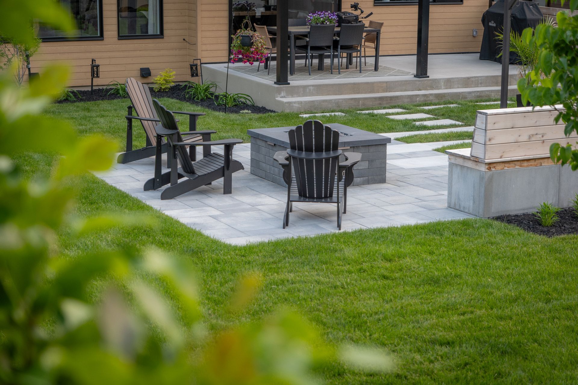 Une terrasse en pierre avec un foyer, trois chaises Adirondack et un banc, aménagée dans une cour arrière près d'une maison.
