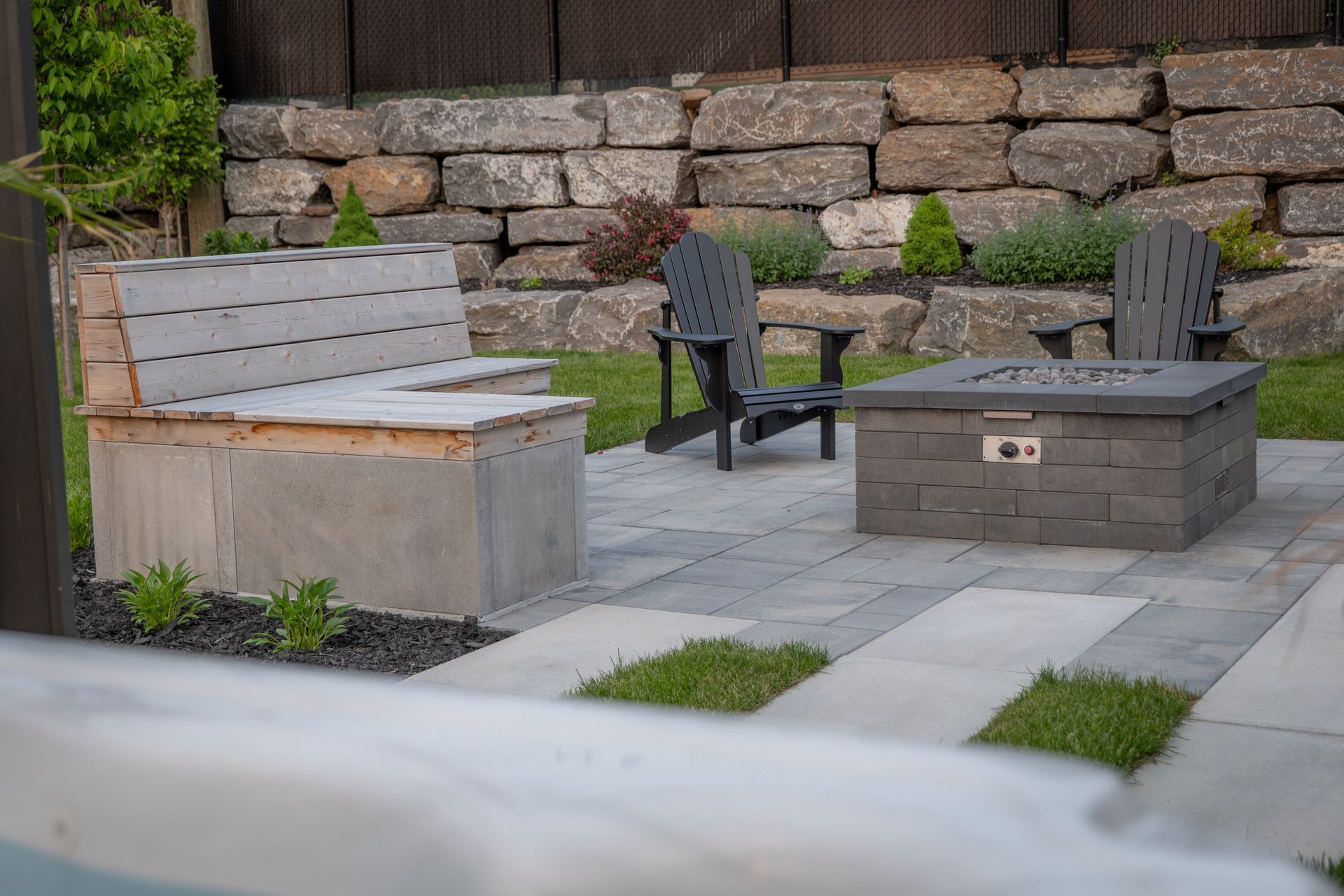 Une terrasse arrière avec un banc en béton en forme de L, deux chaises Adirondack et un foyer carré en pierre sur des dalles de pierre.