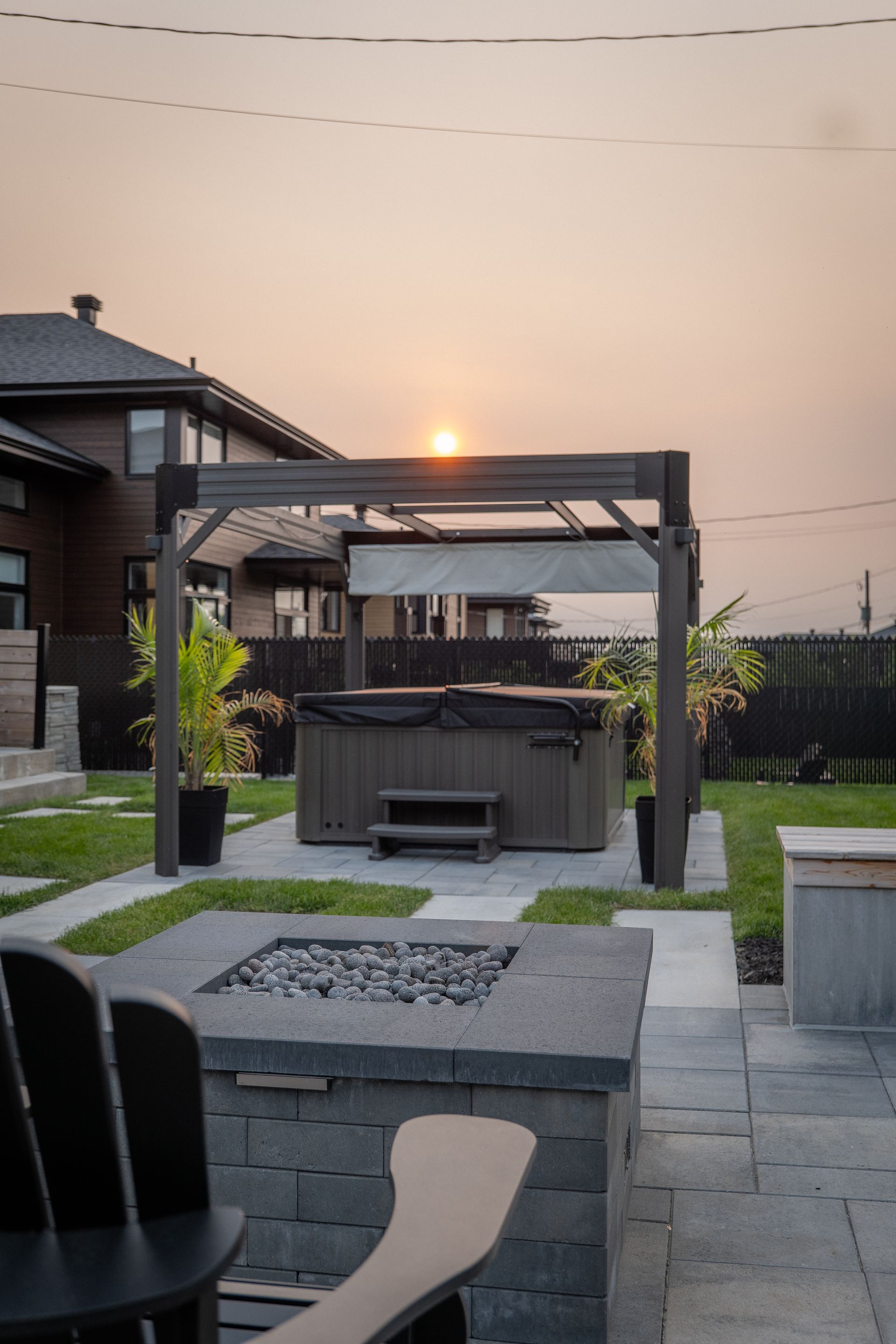 Un jacuzzi sous une pergola dans un jardin au coucher du soleil, avec un foyer au premier plan.