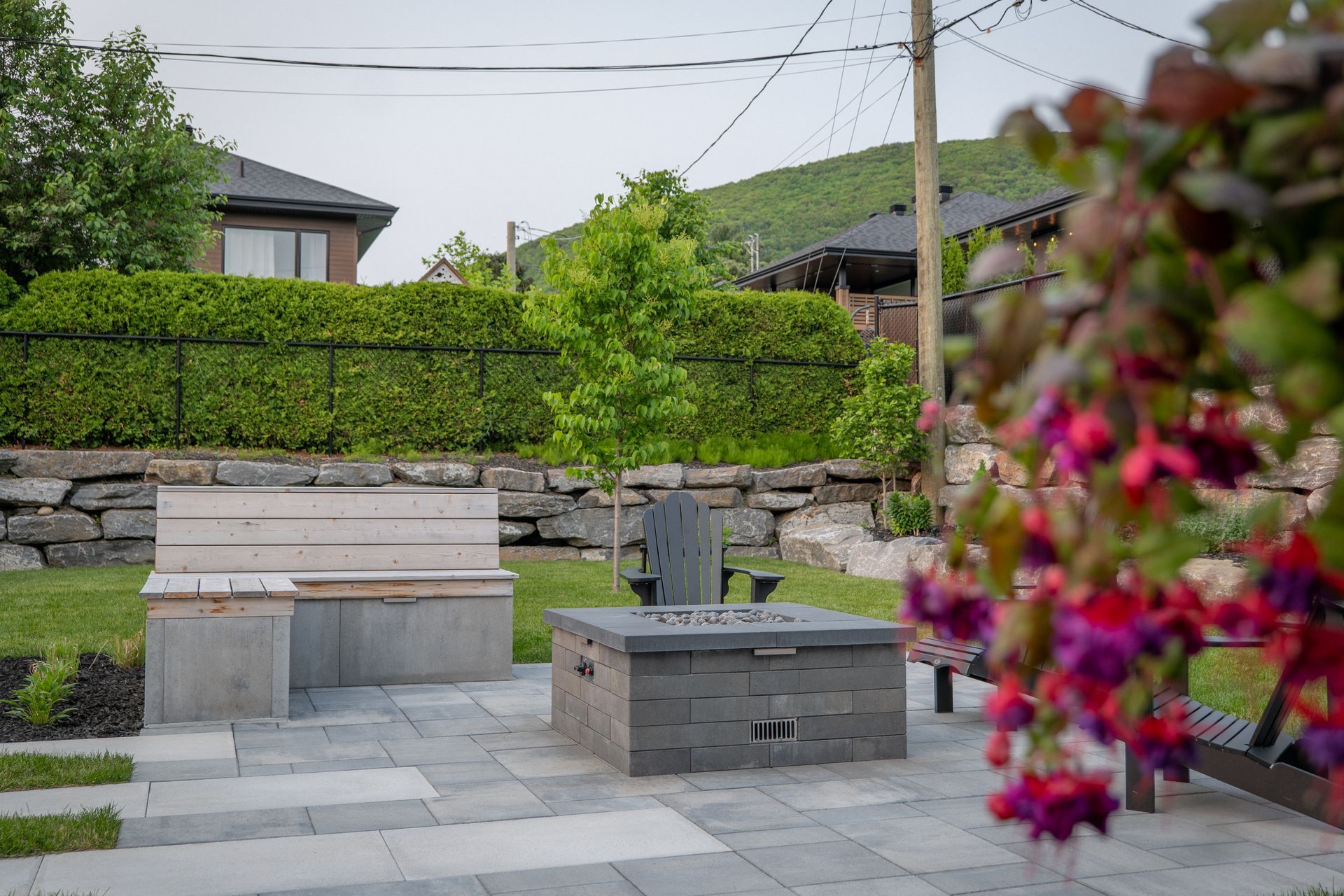 Une terrasse de jardin avec un foyer en pavés gris et un banc en bois clair, adossée à une haie verte et un mur de soutènement en pierre.