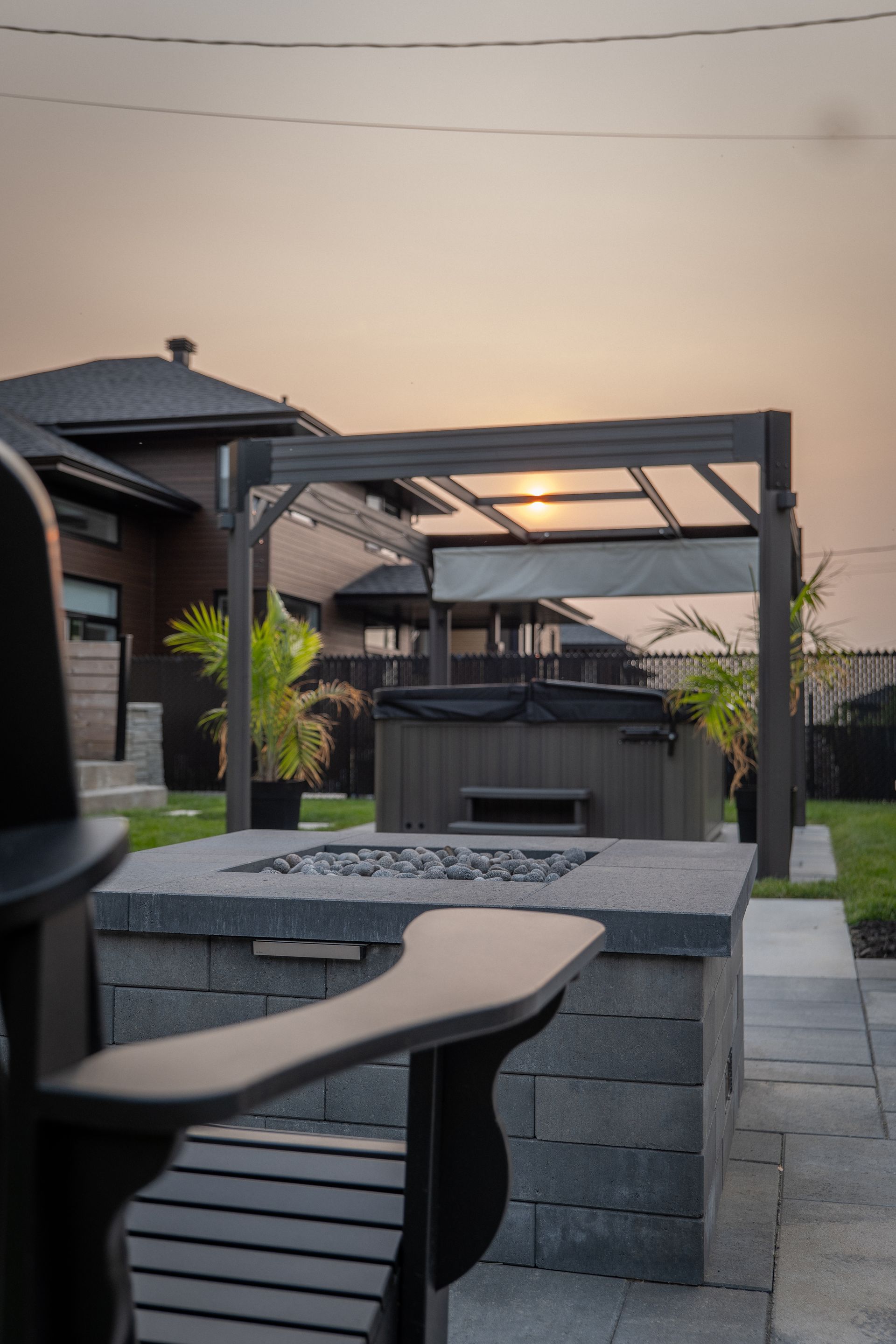 Une terrasse dans un jardin avec un foyer au premier plan, faisant face à un jacuzzi sous une pergola au coucher du soleil.