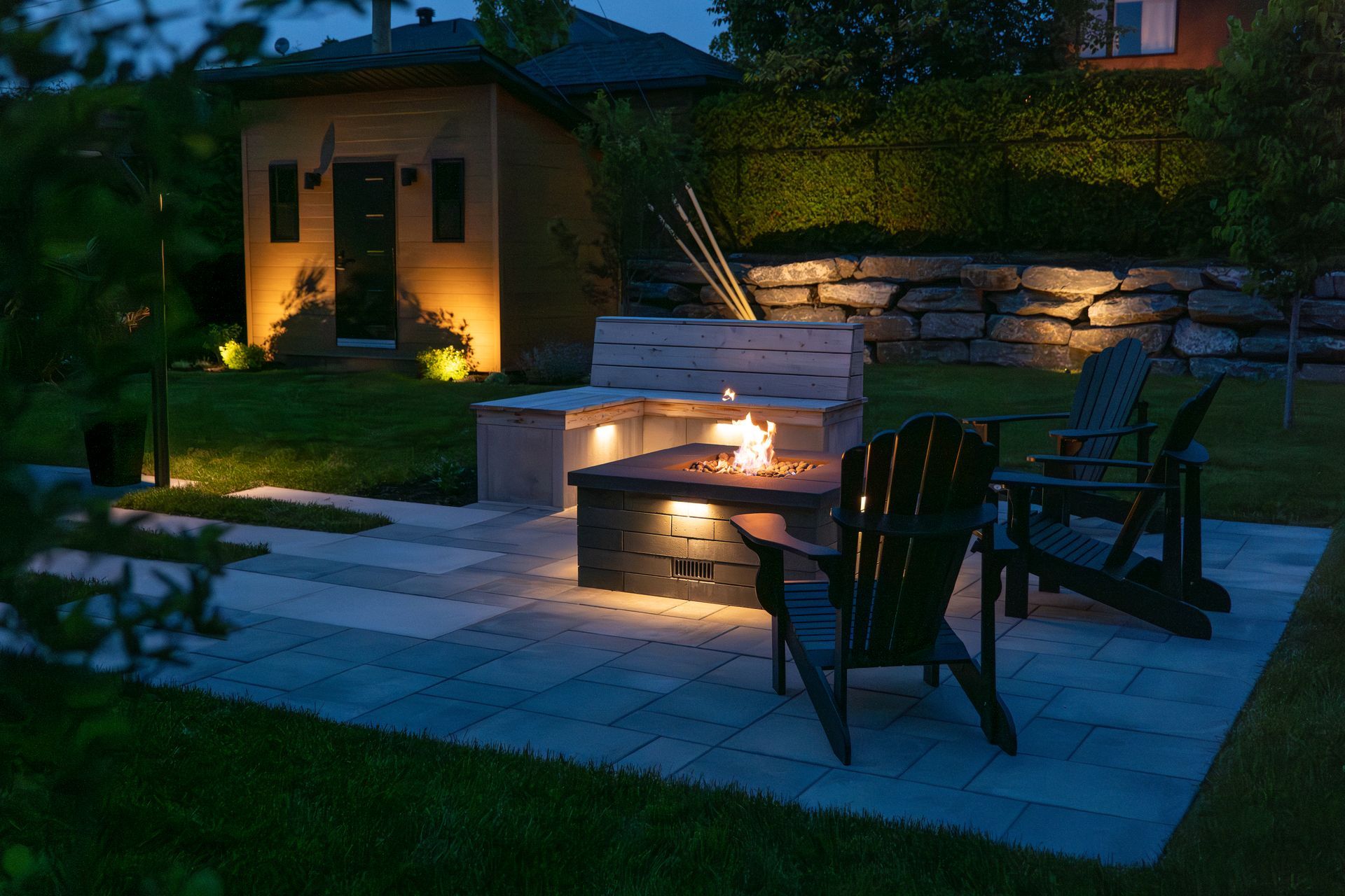 Une terrasse de jardin accueillante au crépuscule, avec un foyer en pierre, des sièges intégrés et deux chaises, le tout éclairé par une lumière chaude.