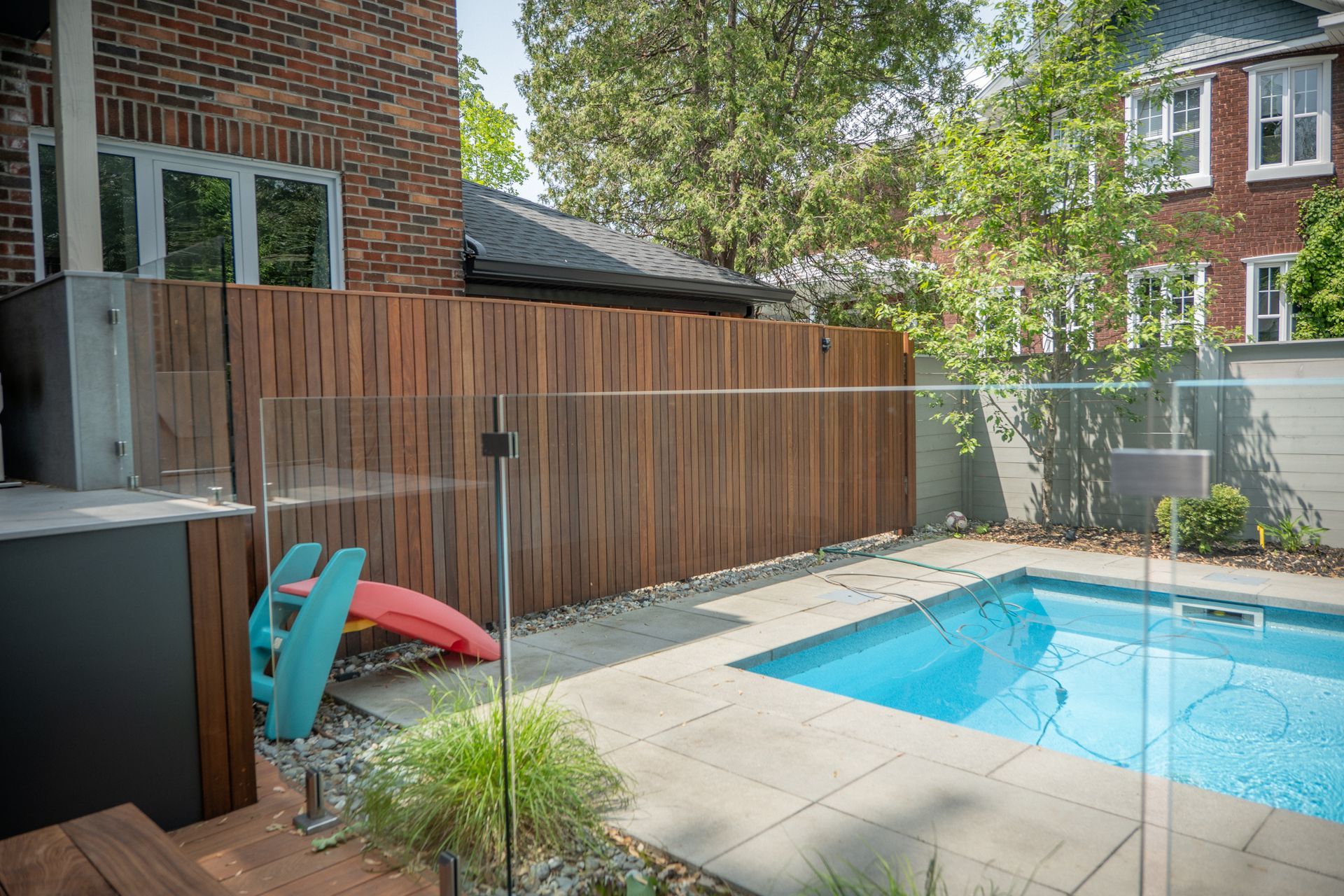 Un jardin avec piscine, terrasse en pierre, clôture en lattes de bois et petit toboggan coloré pour enfants.