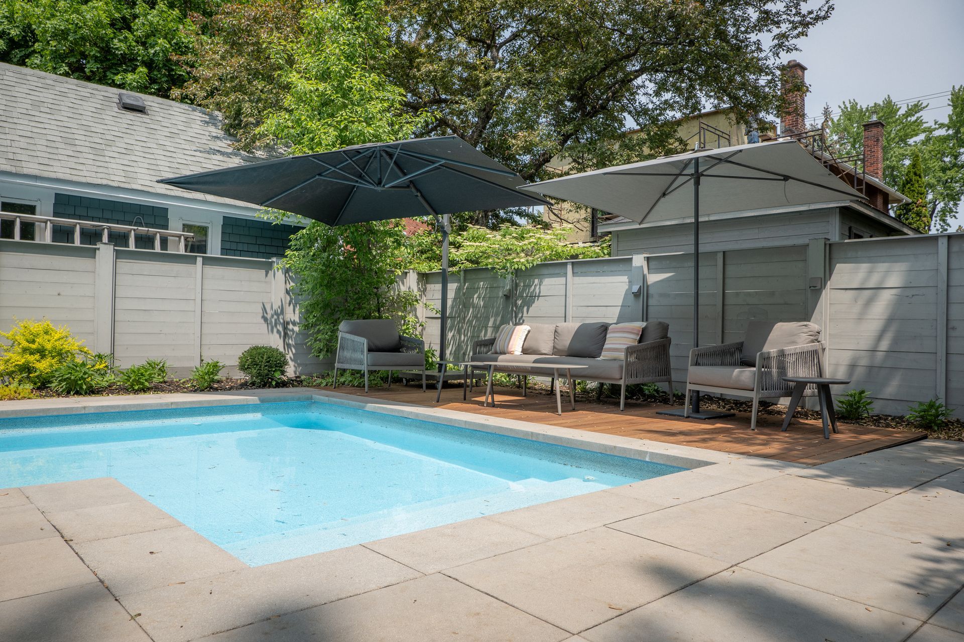 Une piscine turquoise sur une terrasse avec mobilier de jardin, deux parasols et une clôture grise préservant l'intimité dans un jardin.