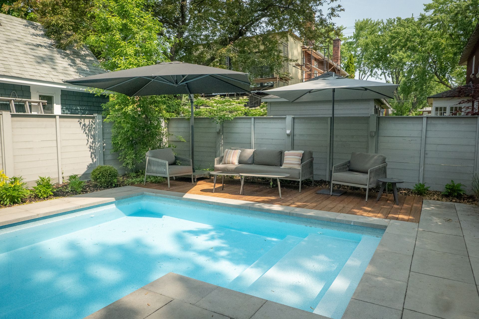 Une piscine dans un jardin avec une eau bleu clair, des meubles de terrasse gris et deux grands parasols adossés à une clôture grise.