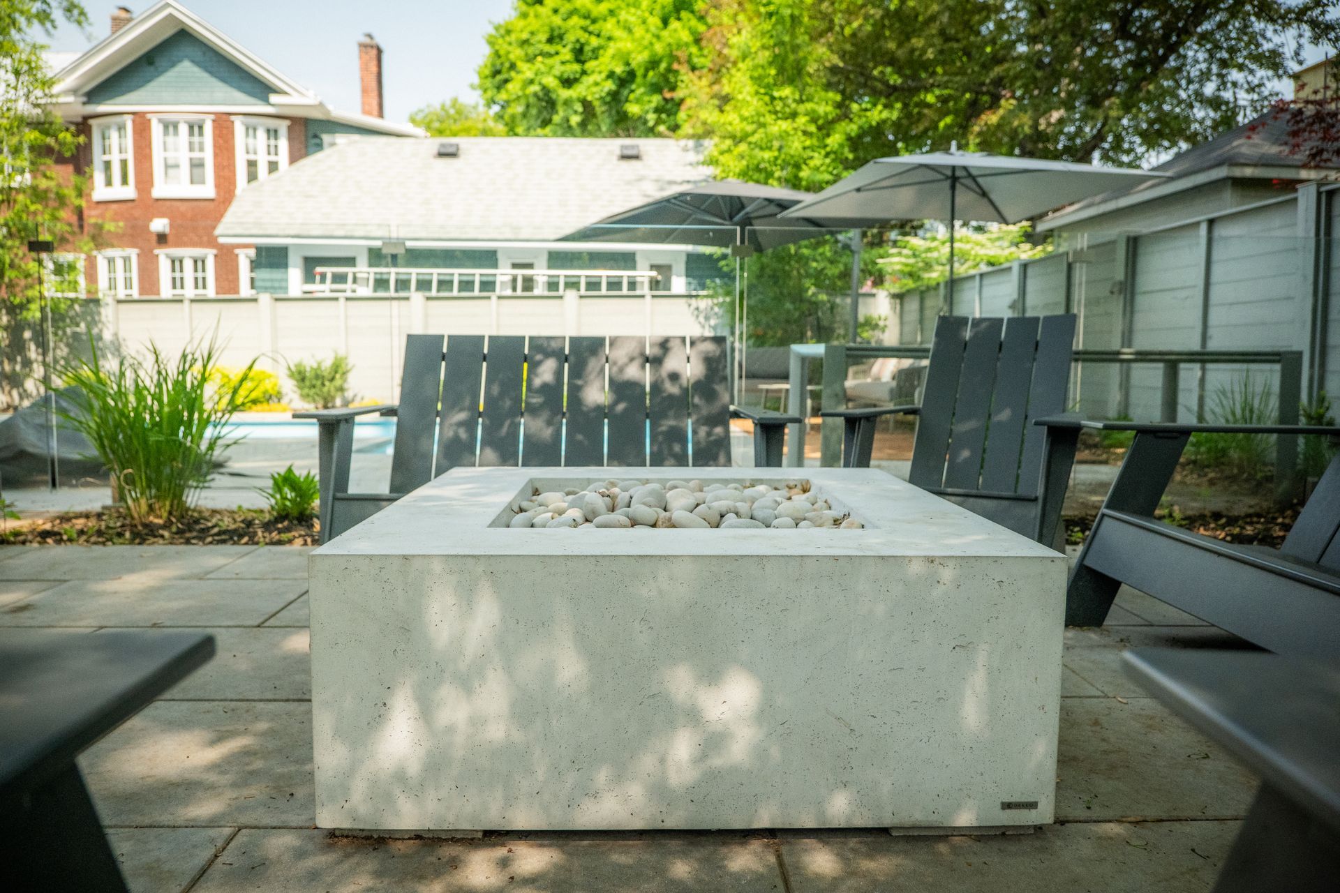 Un foyer carré en béton sur une terrasse en pierre, entouré de chaises sombres, avec des maisons et de la verdure en arrière-plan.
