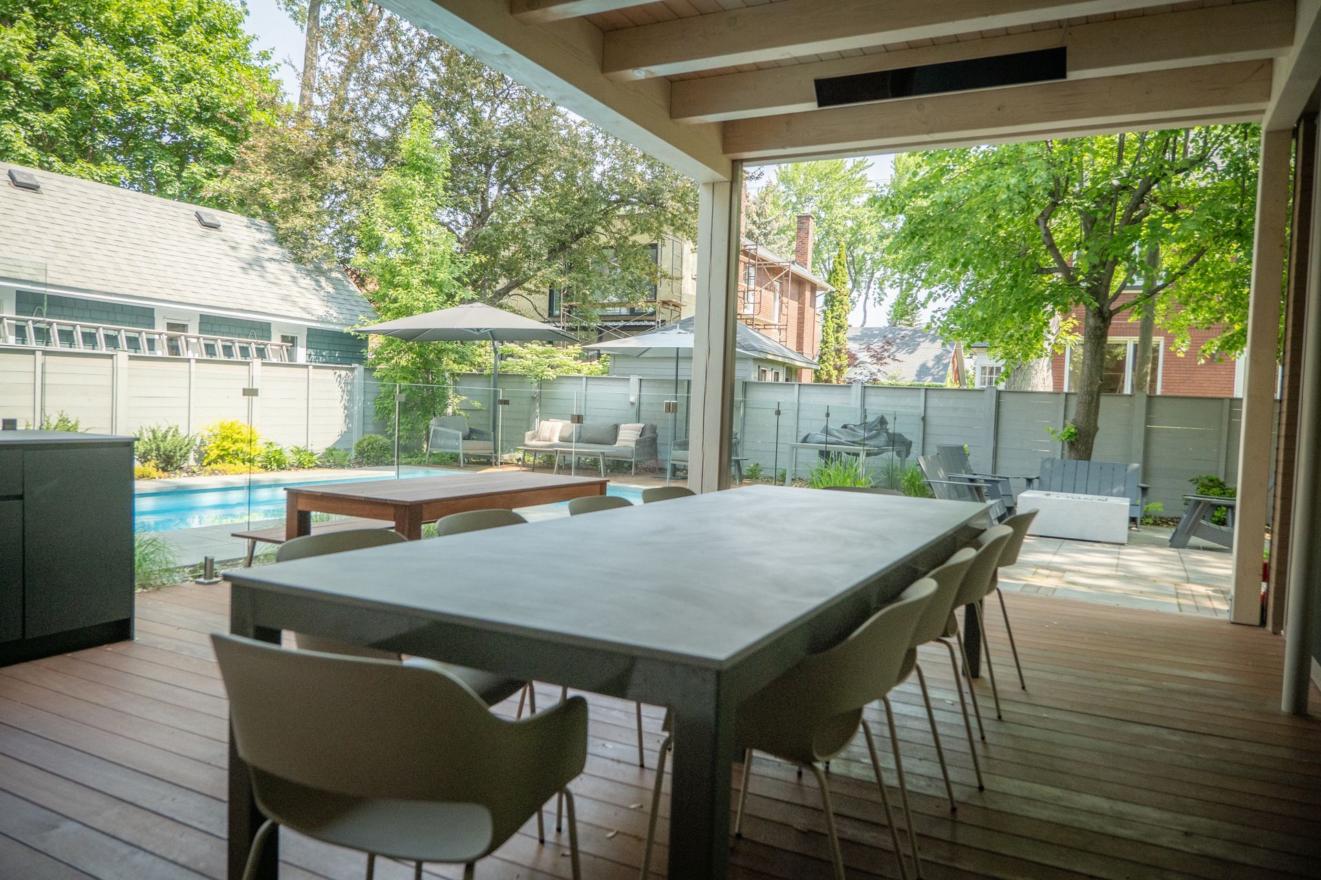 Coin repas extérieur sur une terrasse en bois avec une grande table et des chaises grises, donnant sur une piscine et un jardin clôturé.