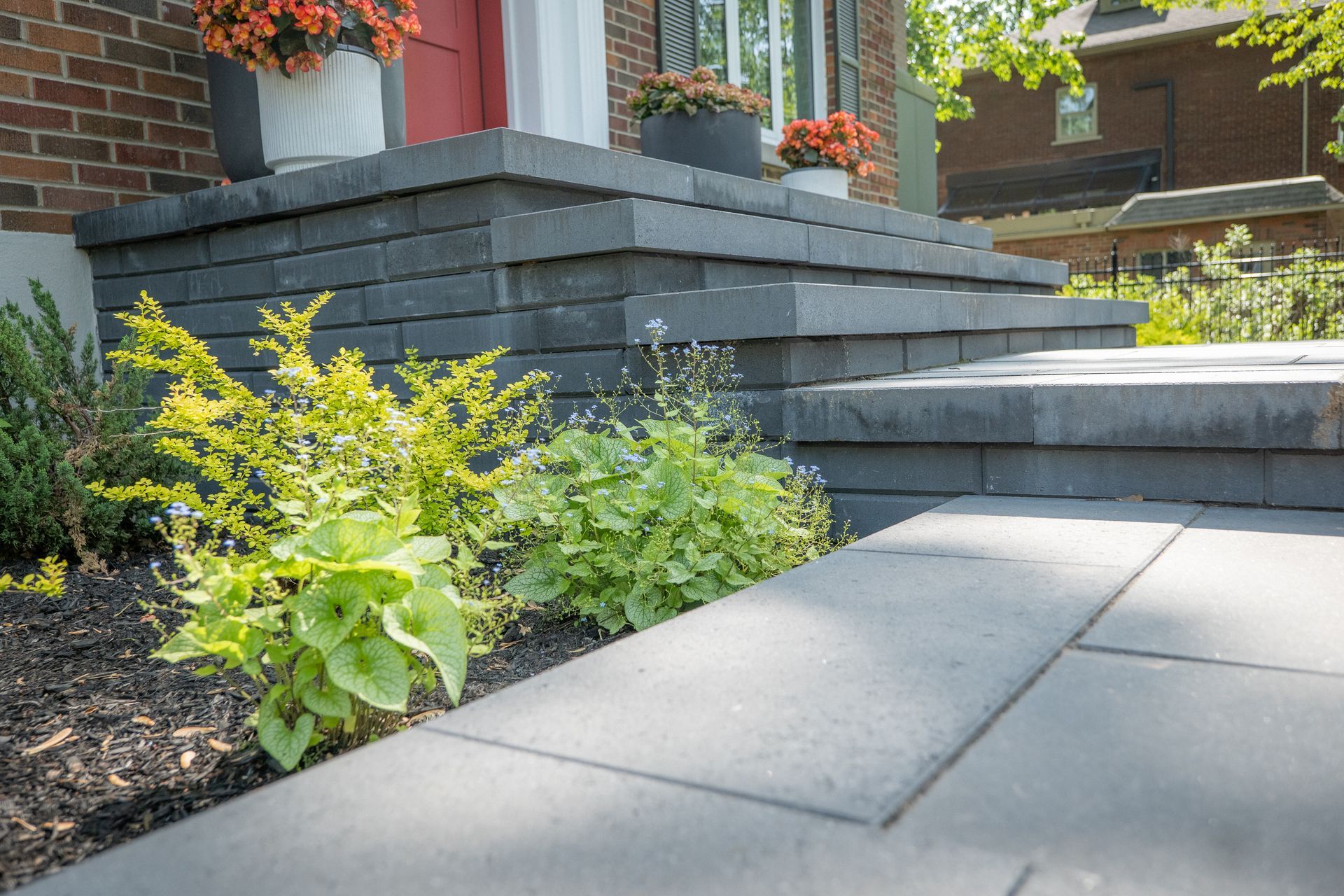 Des marches en pierre grise mènent à l'entrée d'une maison en briques, avec des fleurs en pot et des arbustes de jardin au premier plan.