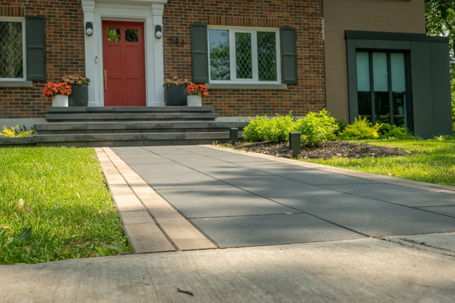 Un chemin pavé mène à l'entrée principale d'une maison en briques avec une porte rouge vif, entourée d'une pelouse et d'aménagements paysagers.