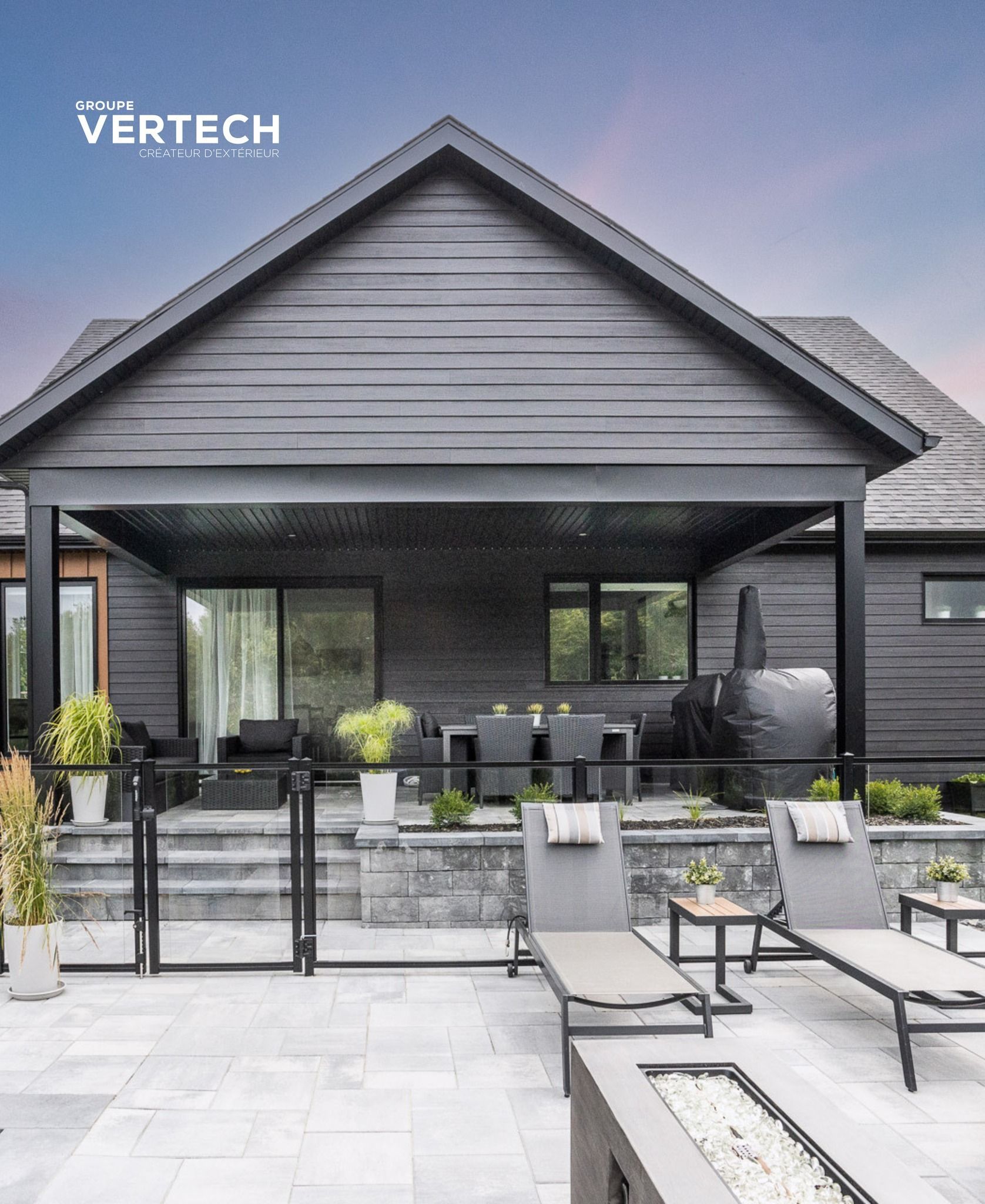 Maison moderne aux façades sombres, avec patio couvert, coin repas extérieur, chaises longues et foyer sur une terrasse en pierre.