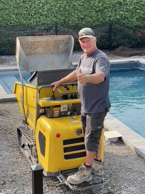 Une personne souriante se tient debout sur un camion-benne jaune à chenilles, le pouce levé, à côté d'une piscine dans un jardin.