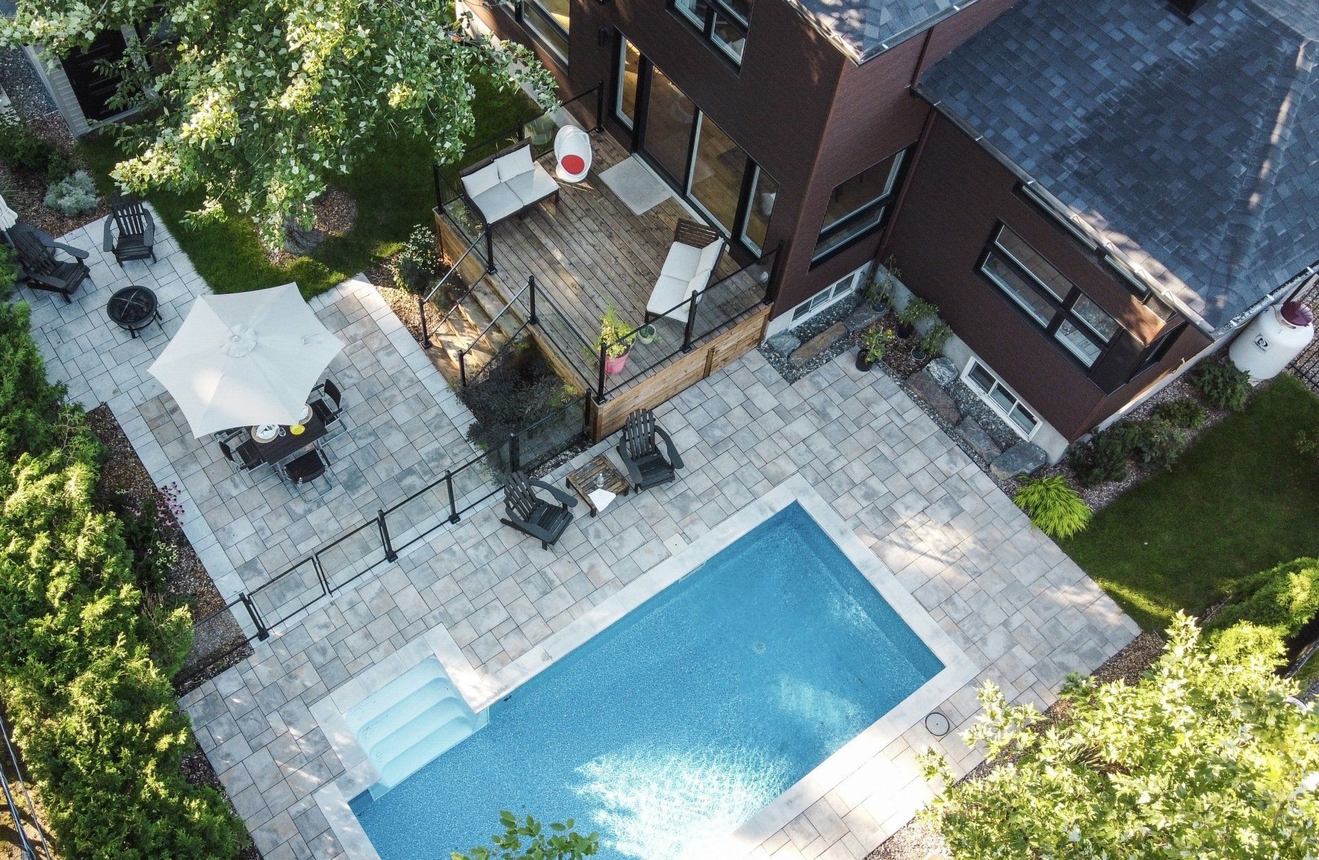 Vue aérienne d'un jardin avec une piscine rectangulaire, une terrasse en pierre, une terrasse en bois et du mobilier de jardin.