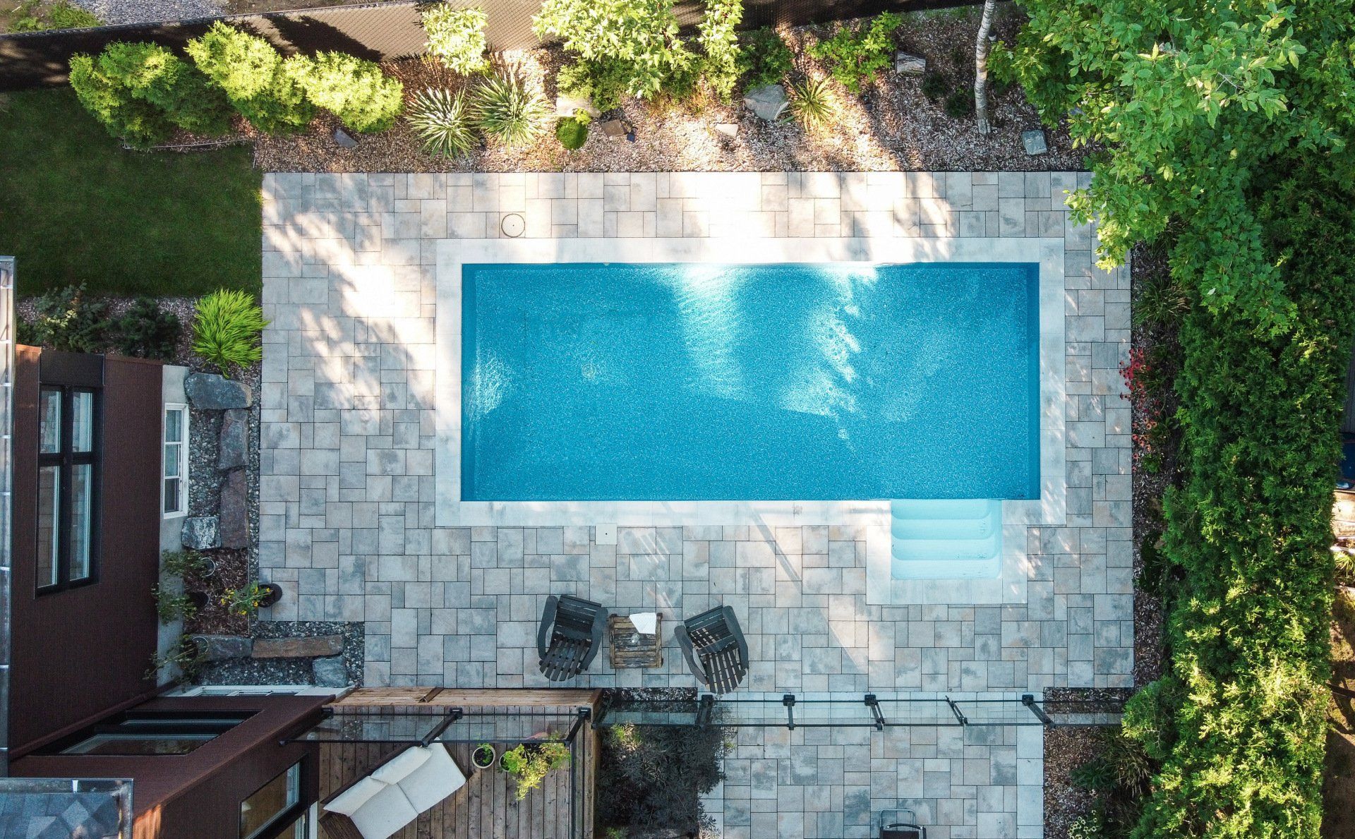 Vue aérienne d'une piscine rectangulaire d'un bleu vif, aménagée sur une terrasse en pierre avec des sièges extérieurs, à proximité d'une maison et d'arbres.