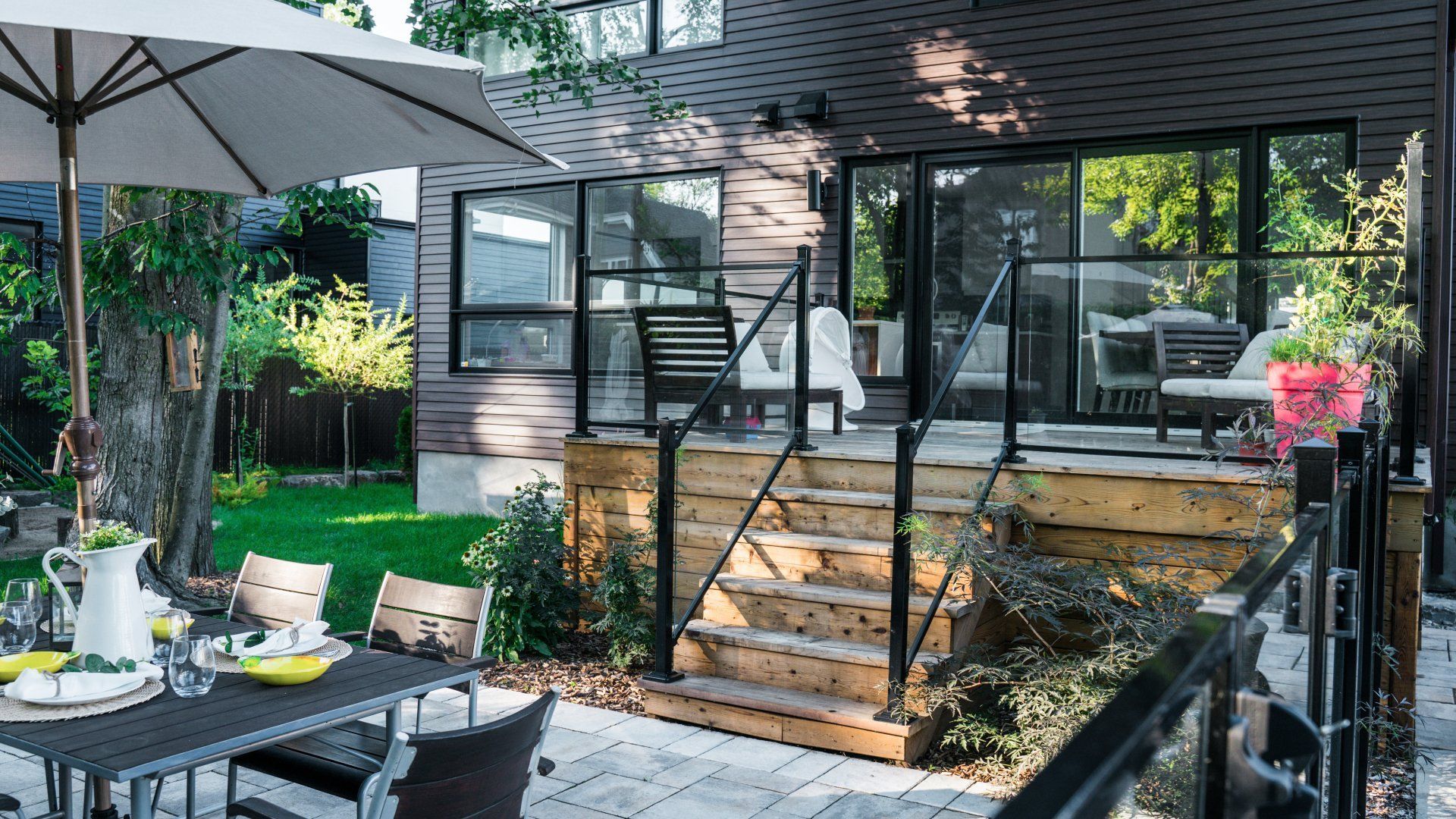 Une terrasse extérieure comprenant une terrasse en bois avec des balustrades en verre, des marches, une table à manger et une façade de maison moderne.