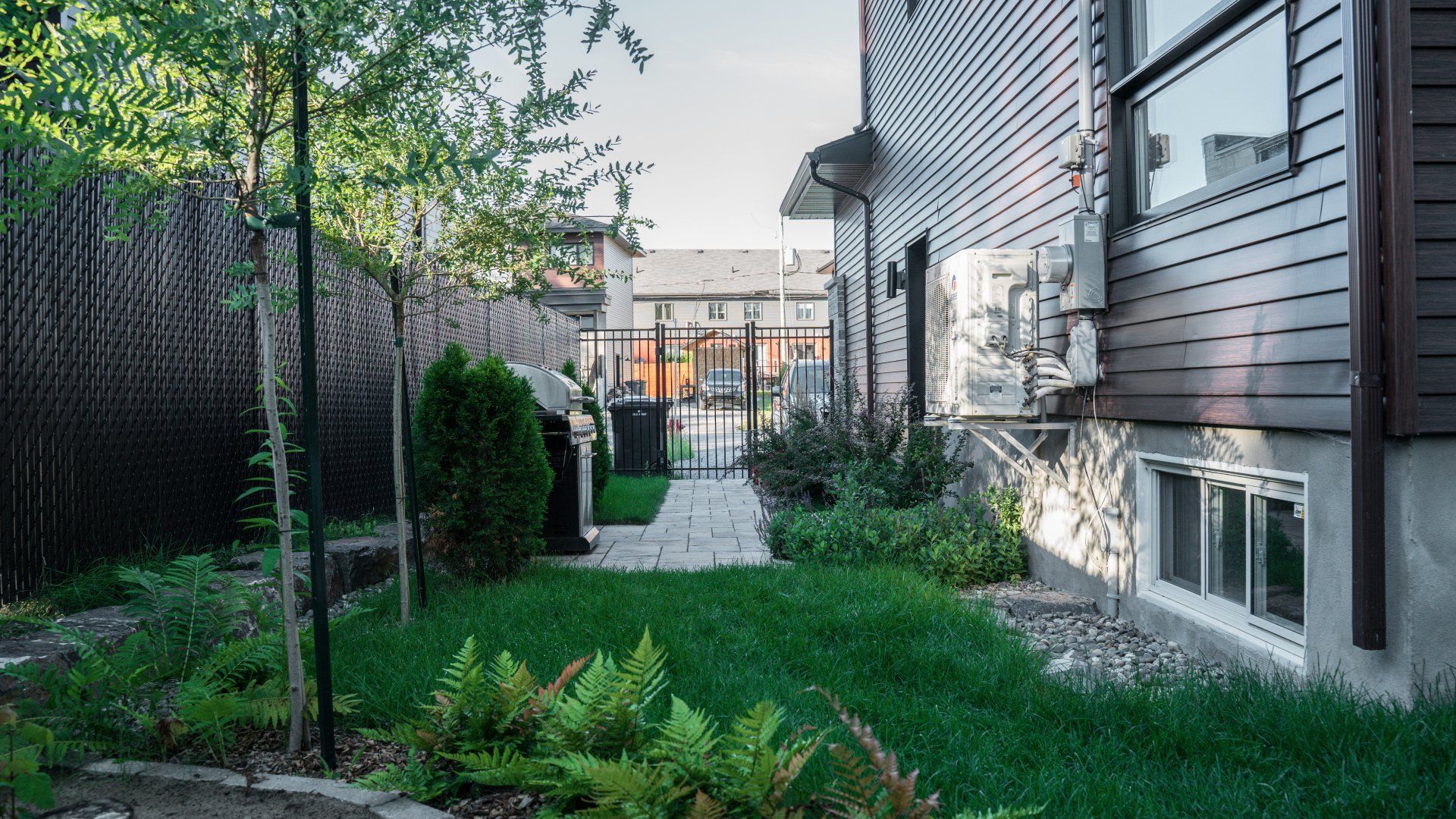 Une cour arrière résidentielle comprenant une pelouse, de petits arbustes, un chemin menant à un portail et une maison grise avec un revêtement extérieur.