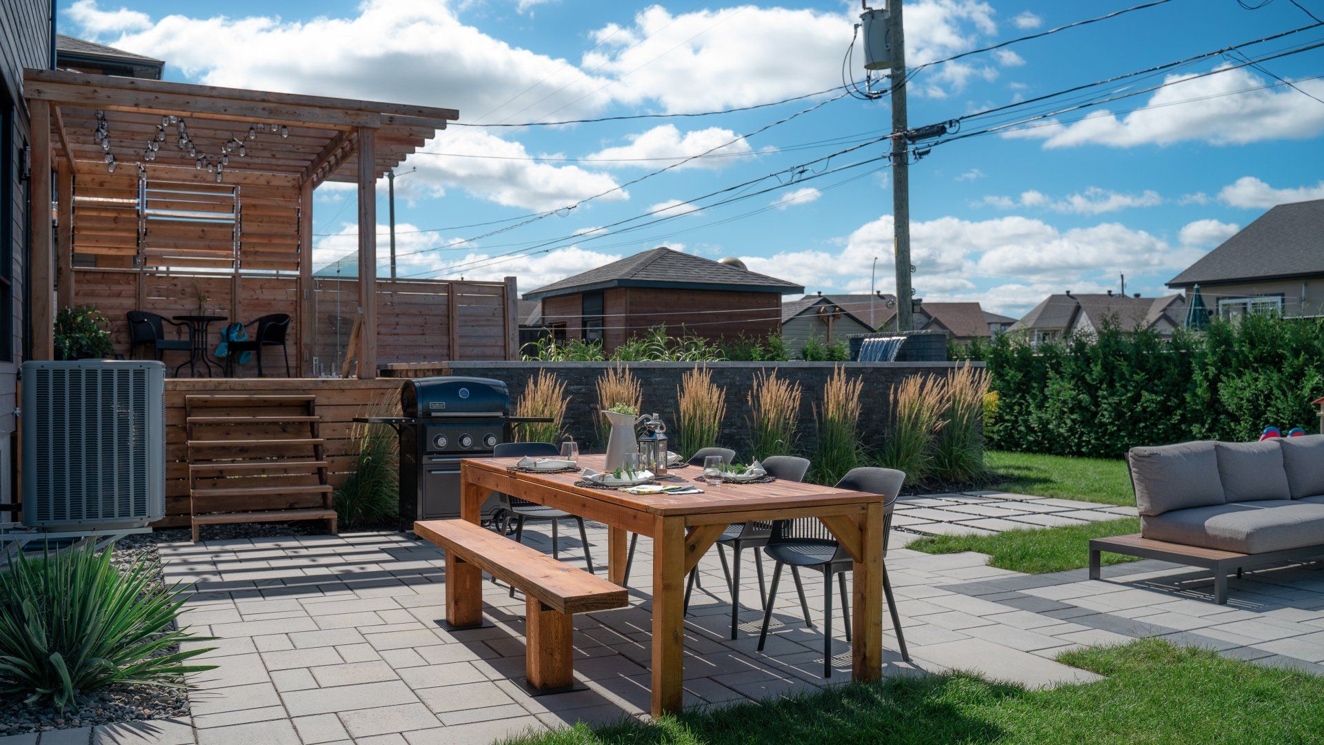 Un canapé d'angle gris foncé et une table basse en bois sont installés sur une terrasse en béton dans un jardin luxuriant et verdoyant.