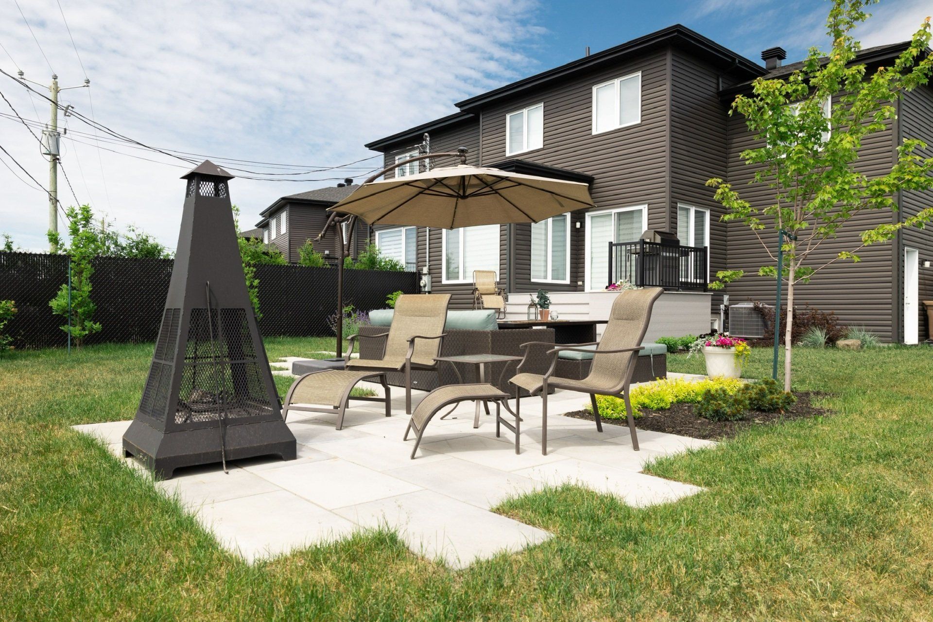 Une terrasse avec un foyer, un parasol et des chaises dans une cour arrière jouxtant une maison brune.