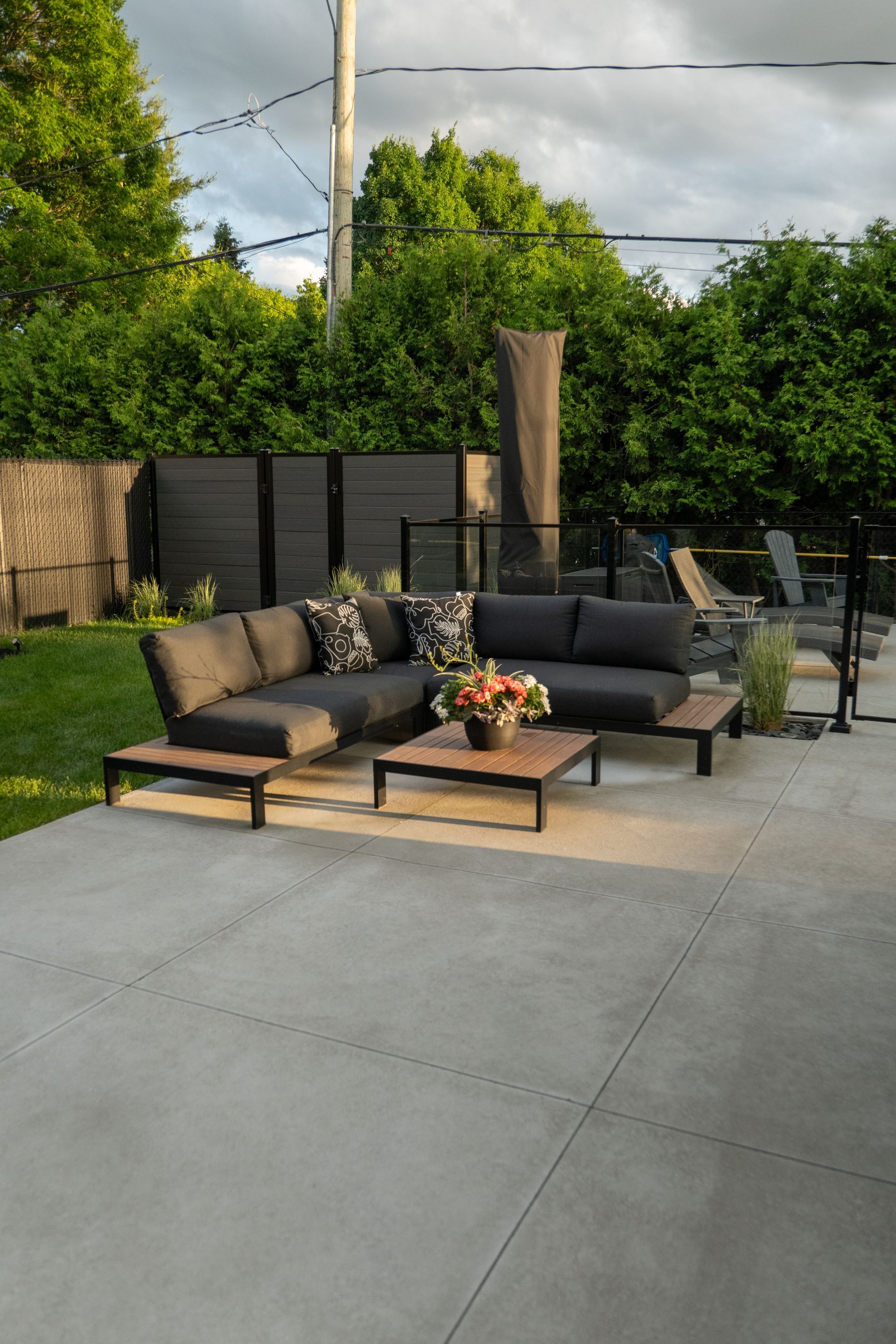 Canapé d'angle gris moderne avec table basse sur une terrasse en béton dans un jardin luxuriant.