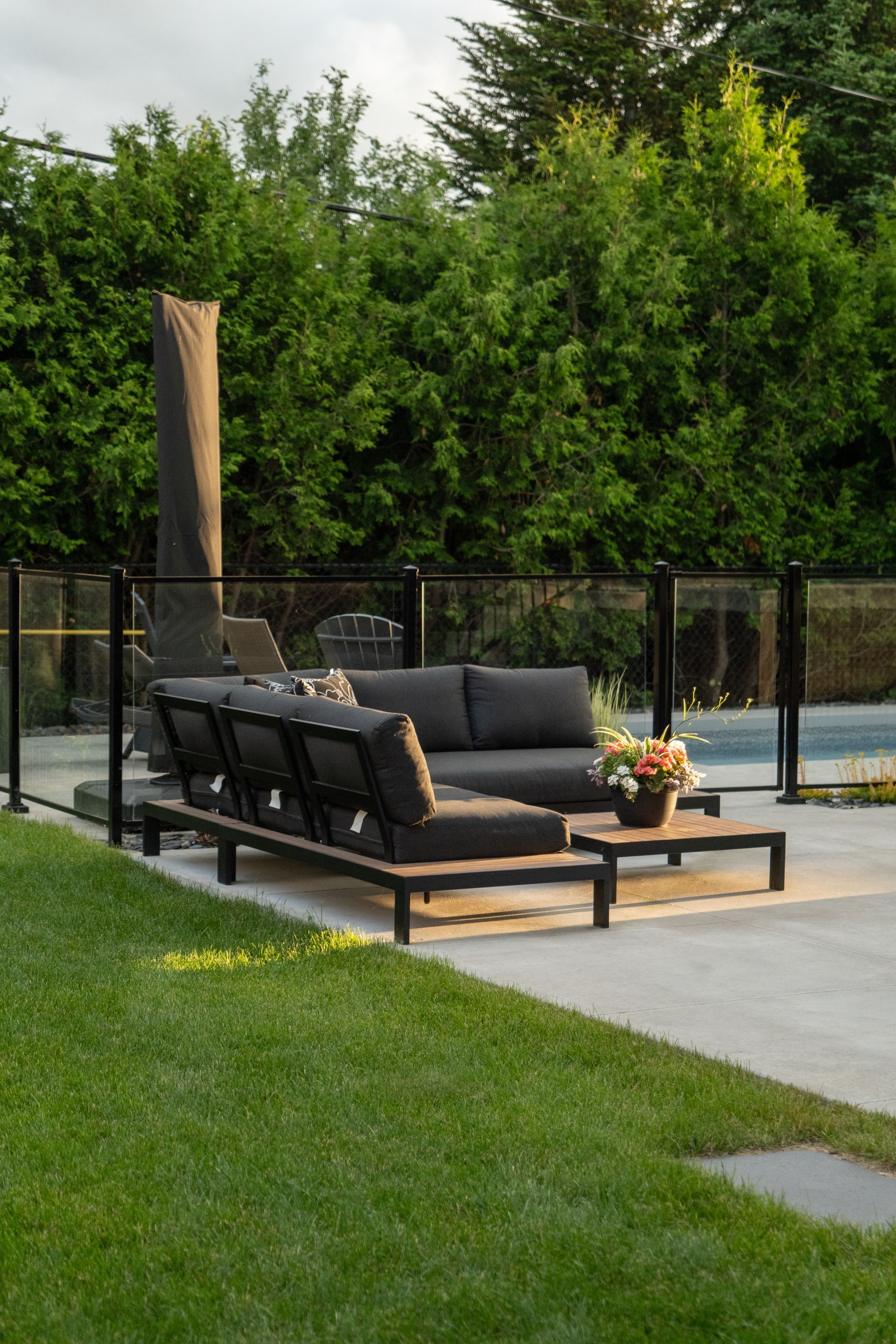 Canapé d'angle d'extérieur gris moderne et table basse assortie sur une terrasse en béton à côté d'une piscine avec une clôture en verre.