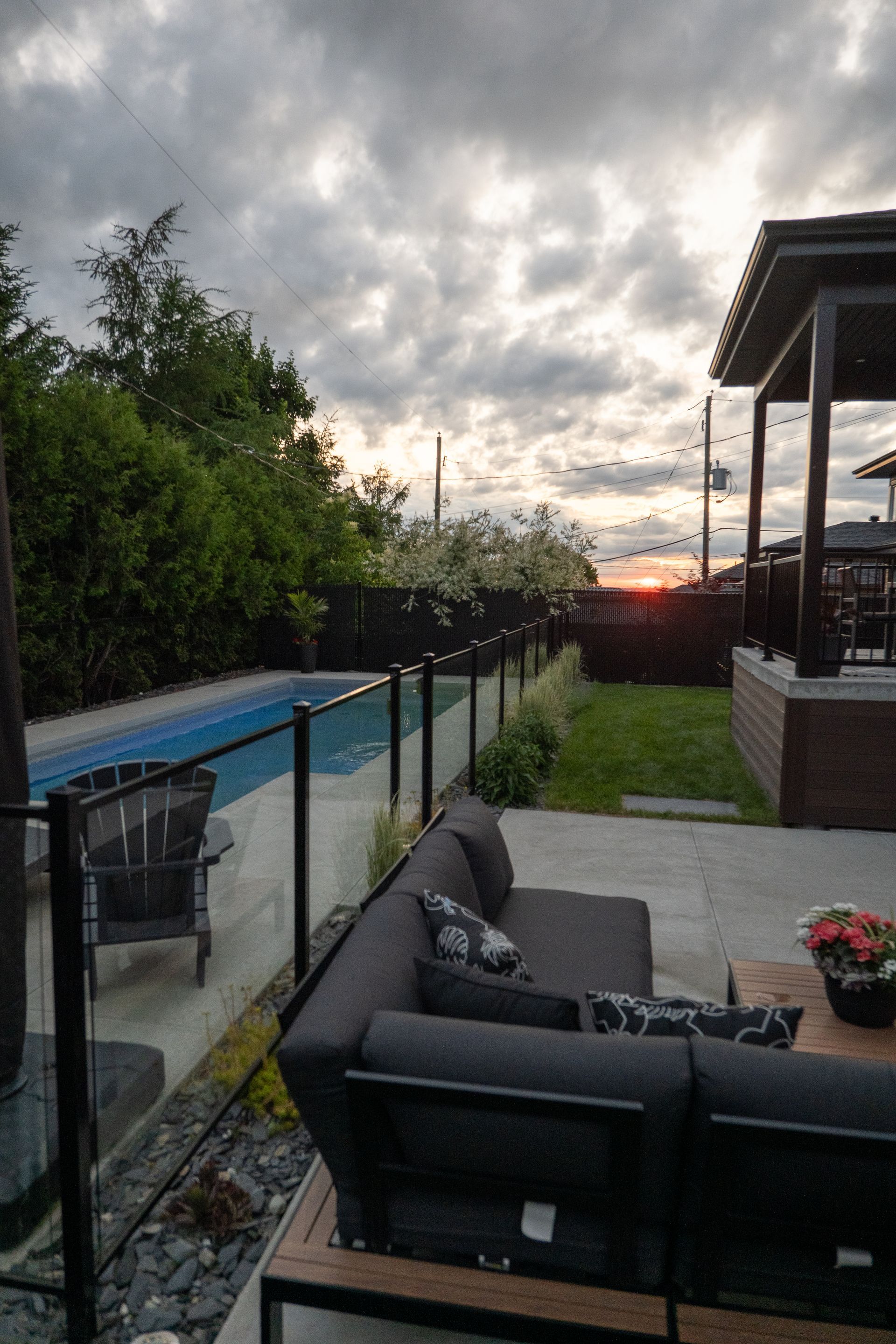 Une terrasse moderne avec un canapé d'angle gris foncé donnant sur une piscine et une pelouse au coucher du soleil sous un ciel nuageux.