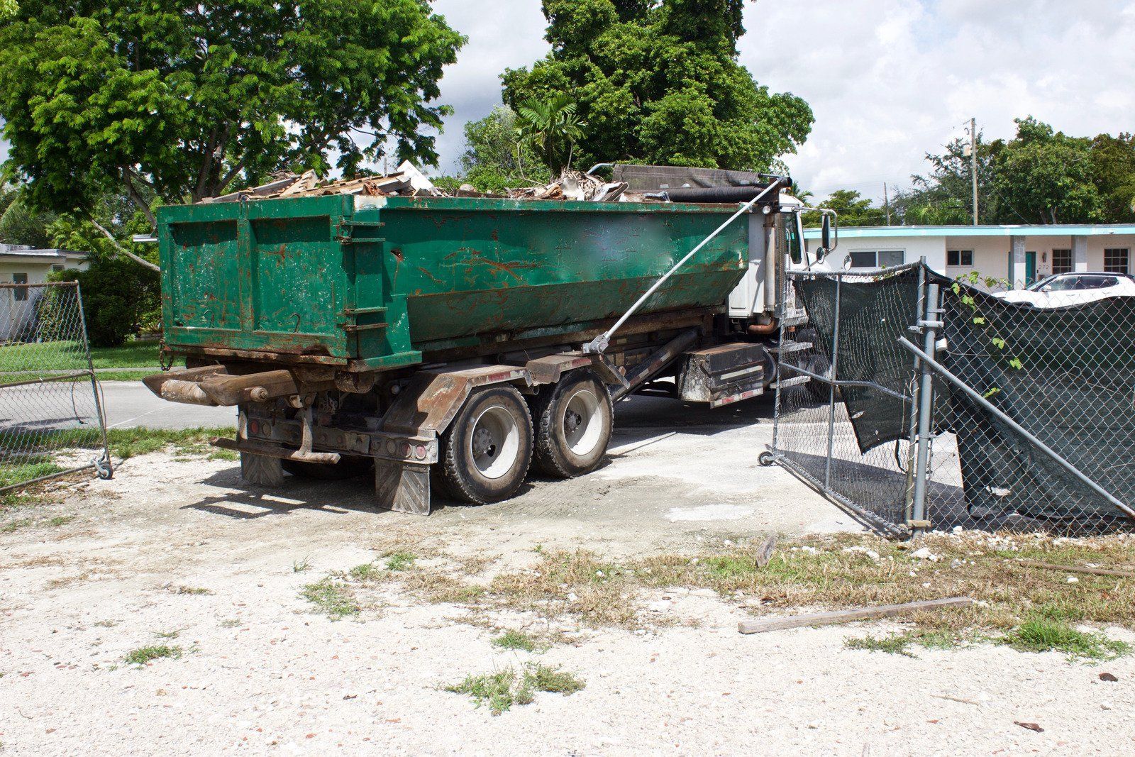 Green dumpster on a truck, filled with debris, parked near a fence and buildings on a gravel surface.
