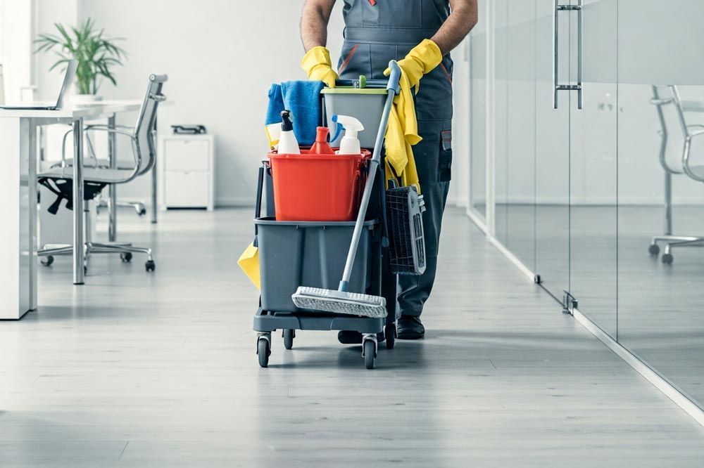 A janitor is cleaning an office with a trolley filled with cleaning supplies.
