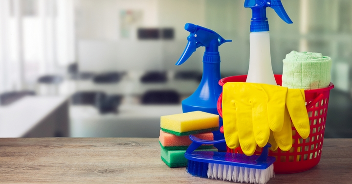 Cleaning supplies, including spray bottles, sponges, a brush, and yellow gloves in a red basket, set on a wooden table.